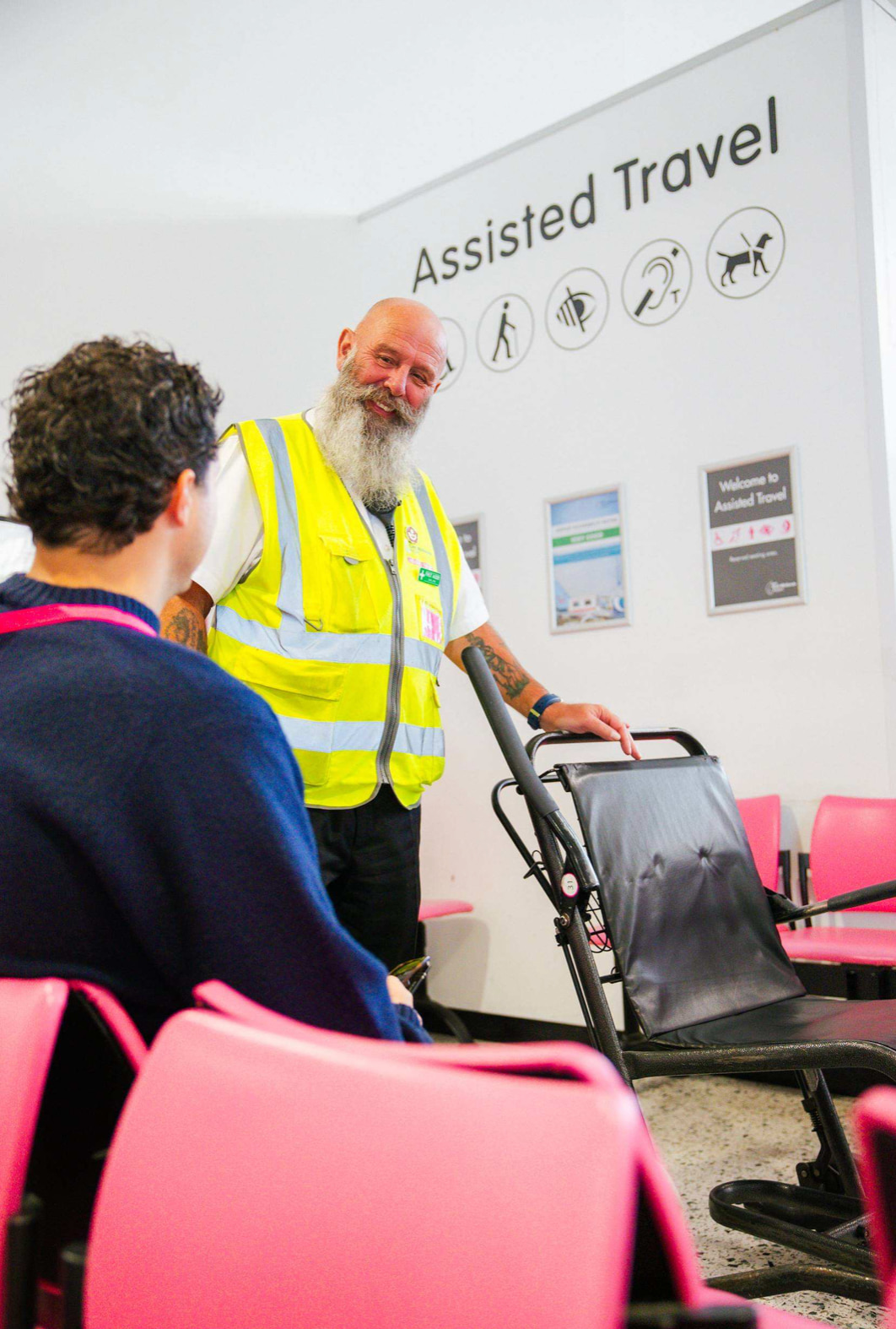 Staff member in a yellow vest assists a traveller with a wheelchair in the "Assisted Travel" area, near pink chairs.