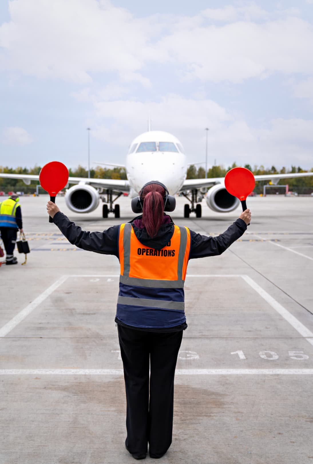 Airport worker guides approaching plane with red paddles, wearing an "Operations" vest and headset on the tarmac.