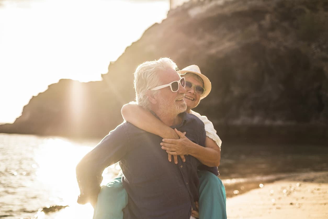 An older couple enjoying a playful moment on a sunlit beach. The man gives the woman a piggyback ride, both smiling and wearing sunglasses.