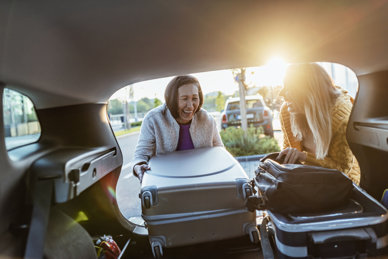 Two people laughing as they load luggage into a car trunk, with sunlight streaming in from the background.