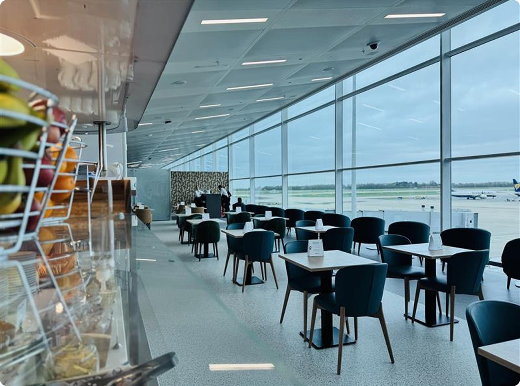 Spacious airport lounge with tables, chairs, and large windows overlooking the runway; fruit basket in the foreground.