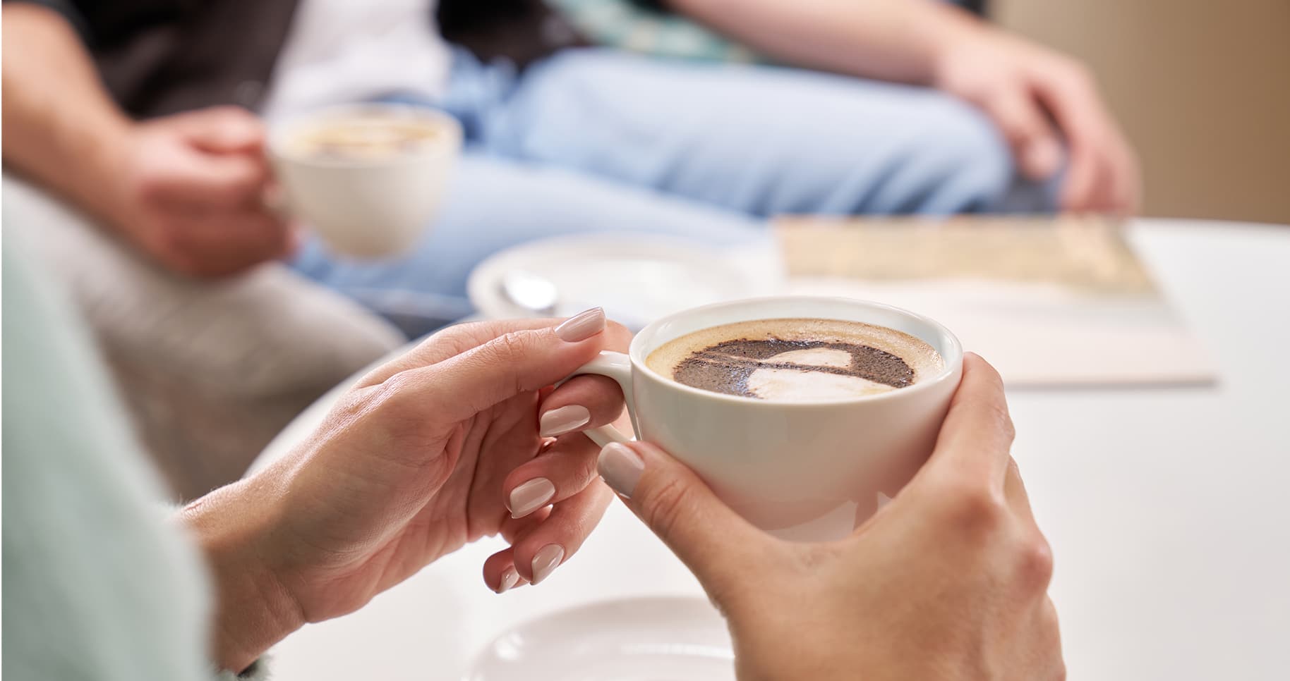 A person holding a cup of coffee with frothy art, sitting at a table with another person drinking coffee in a relaxed setting.