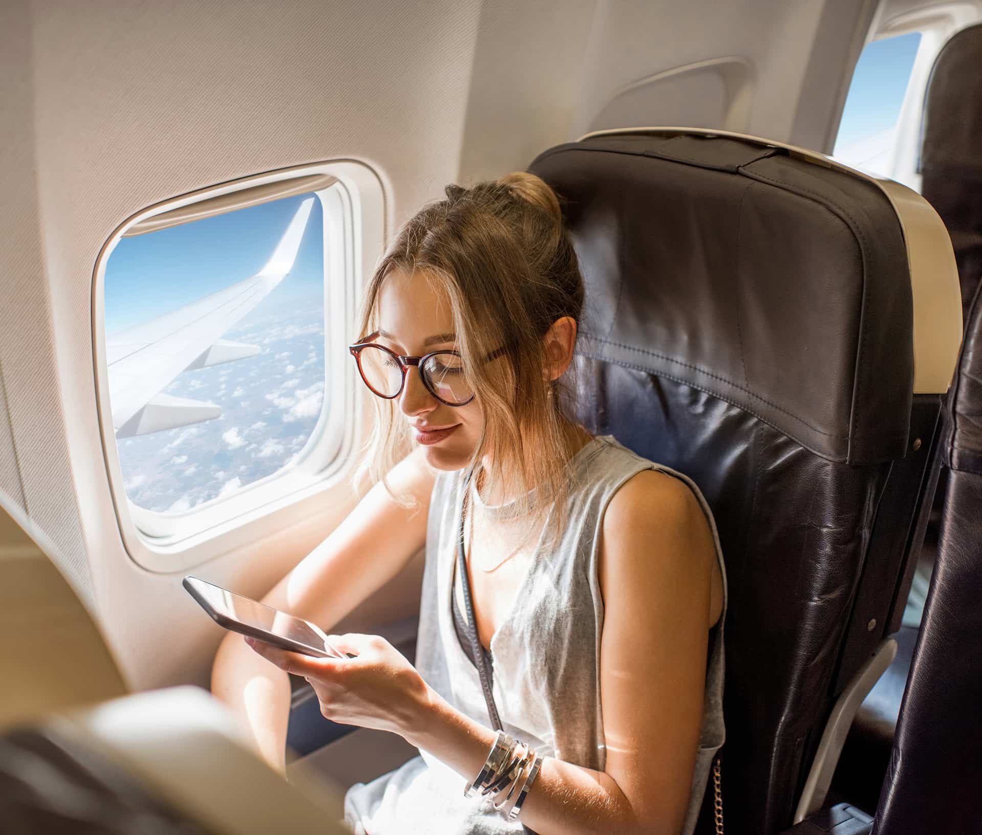 Woman with glasses using a smartphone, seated by a plane window with views of the sky and clouds outside.