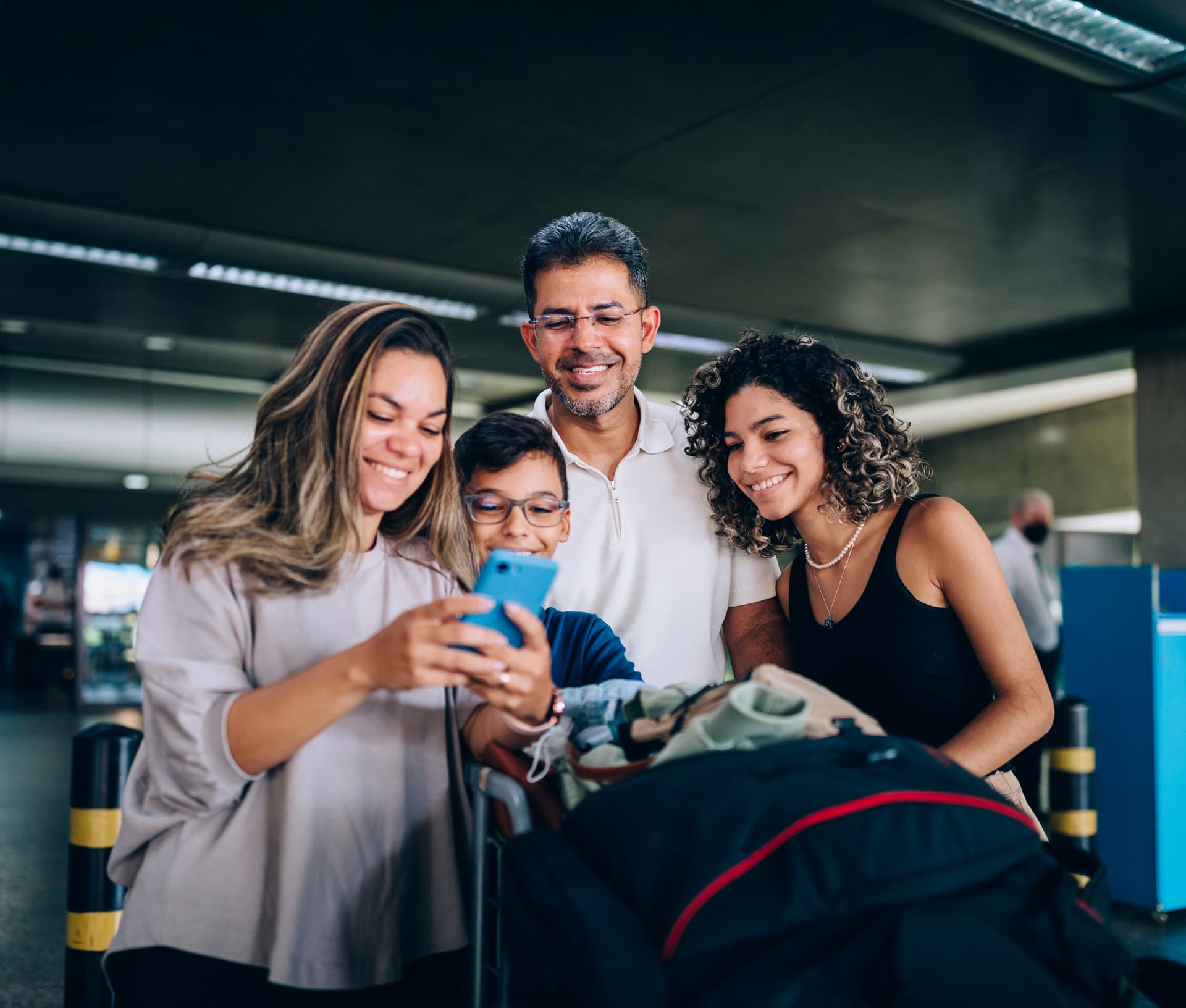 A smiling family at the airport looks at a smartphone. They are gathered around luggage, suggesting they are traveling.