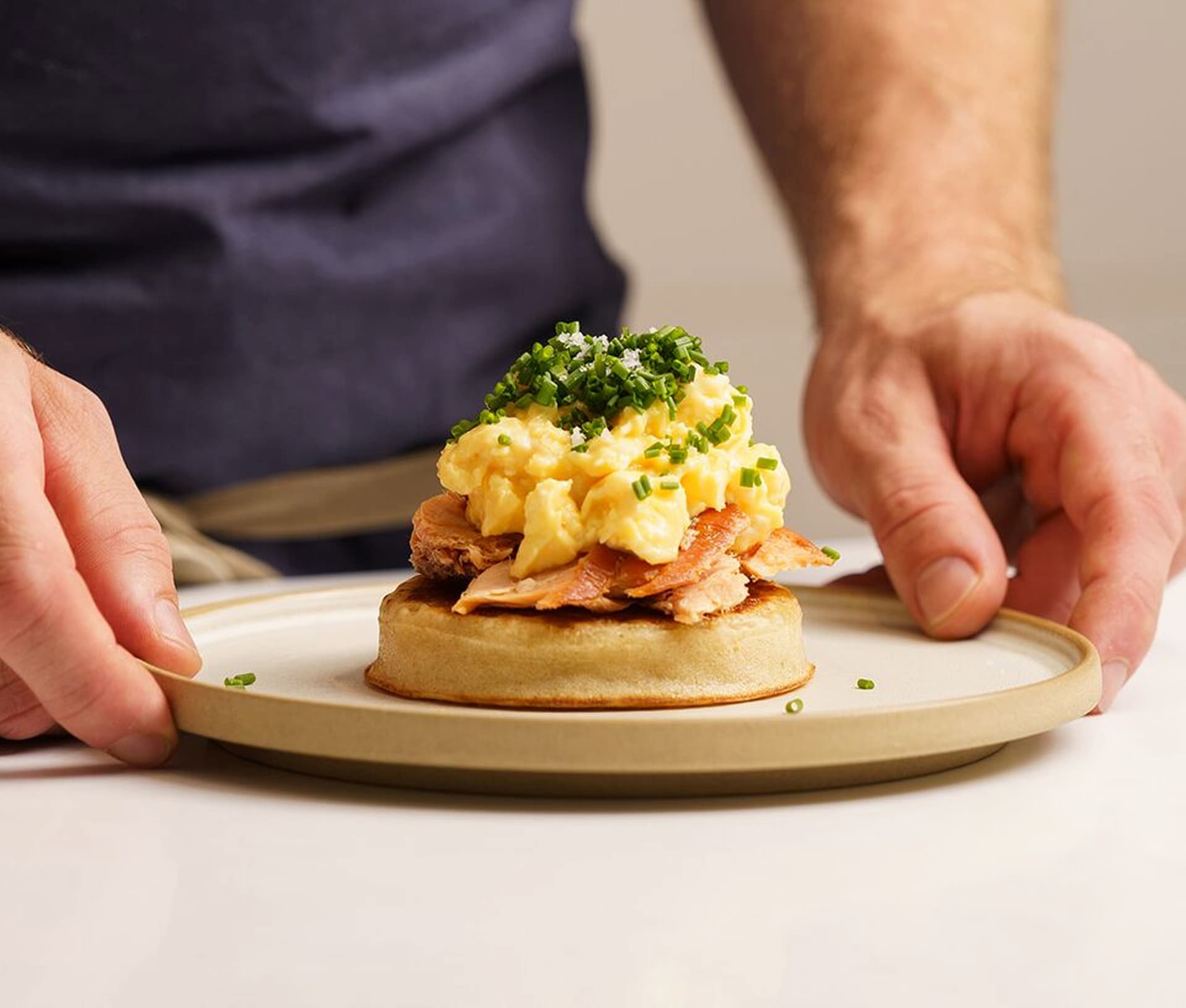 Hands holding a plate with a dish featuring scrambled eggs, bacon, and chopped chives on a round bread base.