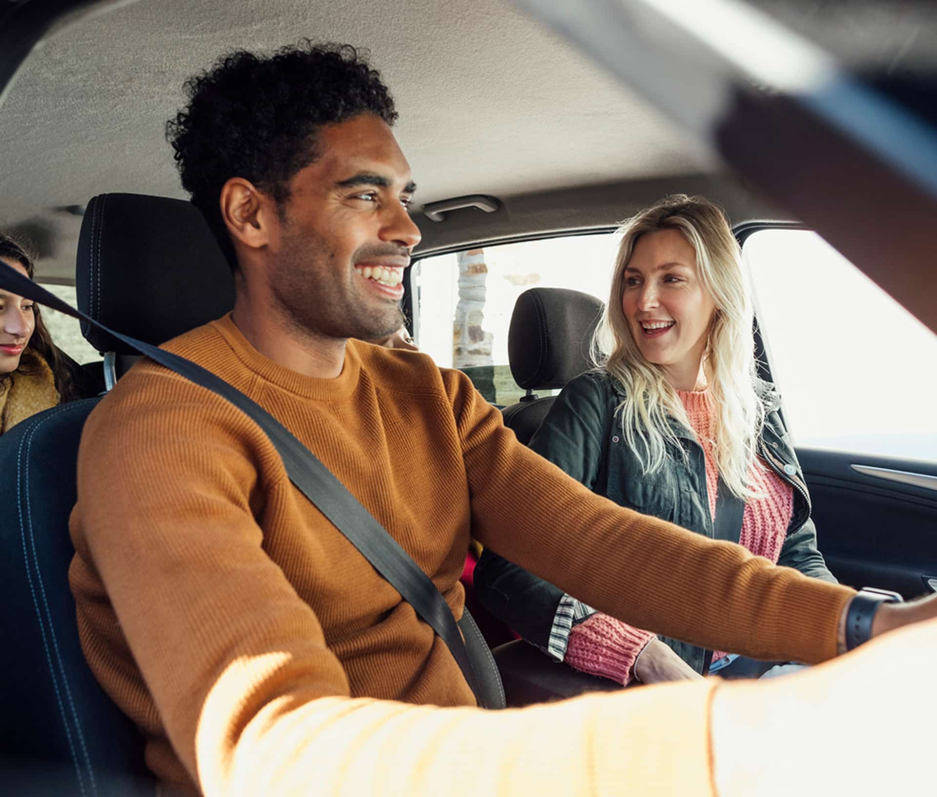 A man driving a car, smiling, with a woman in the passenger seat. They are conversing happily, with another passenger visible in the backseat.