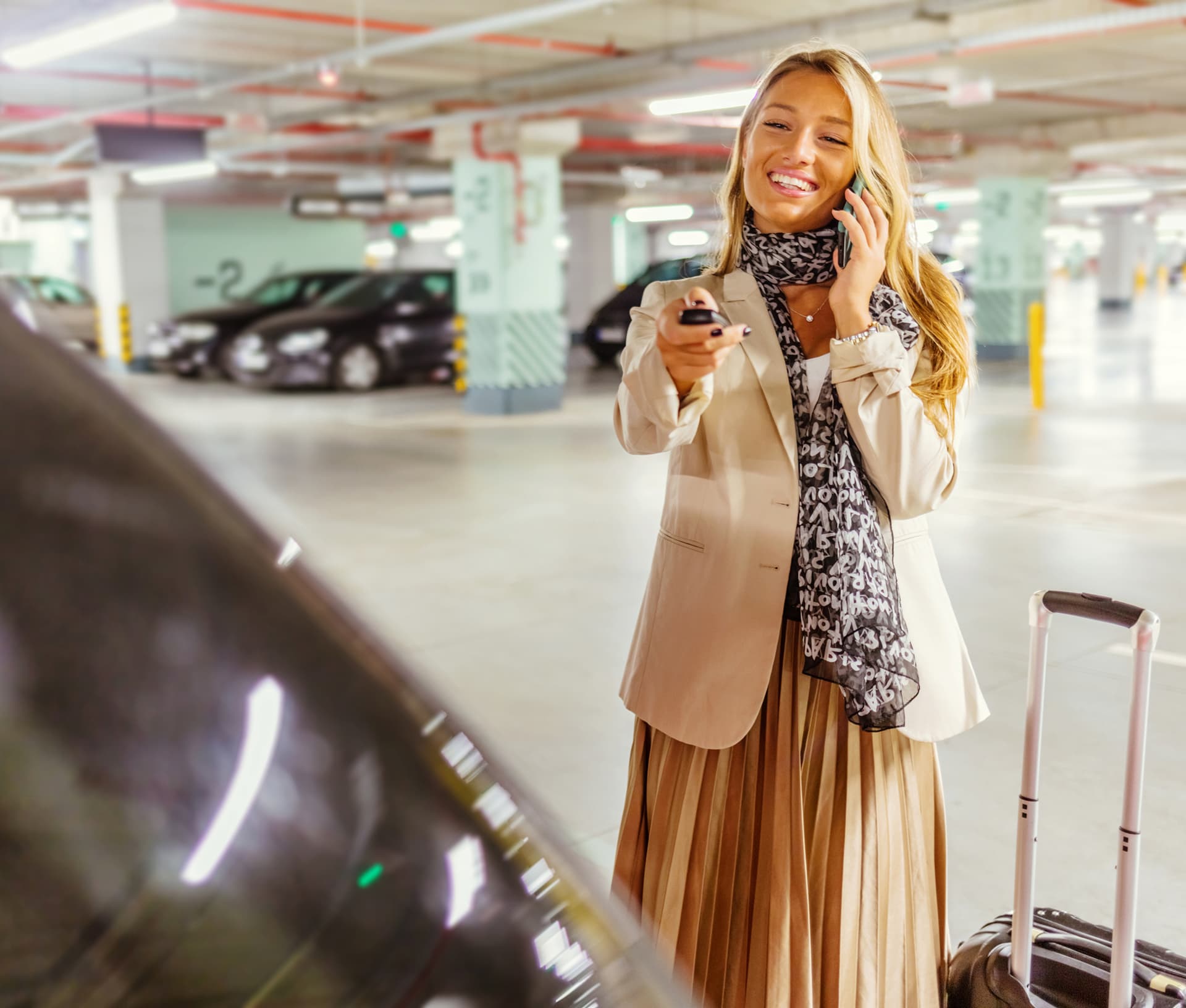 Smiling woman in a beige outfit uses key fob and talks on phone in a parking garage, next to luggage.