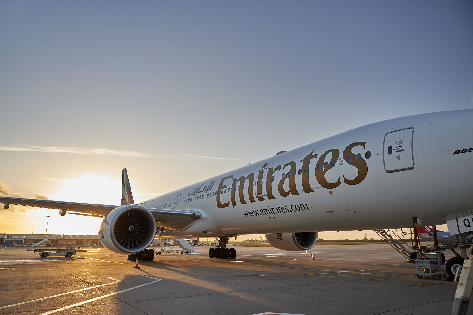 An Emirates airplane is parked on an airport tarmac at sunrise, with its engines and branding visible under a clear sky.