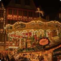 A brightly lit carousel with festive decorations at a bustling night-time fair, surrounded by a crowd and traditional buildings.