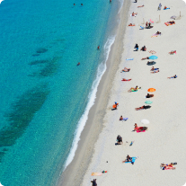 Aerial view of a beach with turquoise water, scattered sunbathers, colorful umbrellas, and gentle waves lapping at the sandy shore.