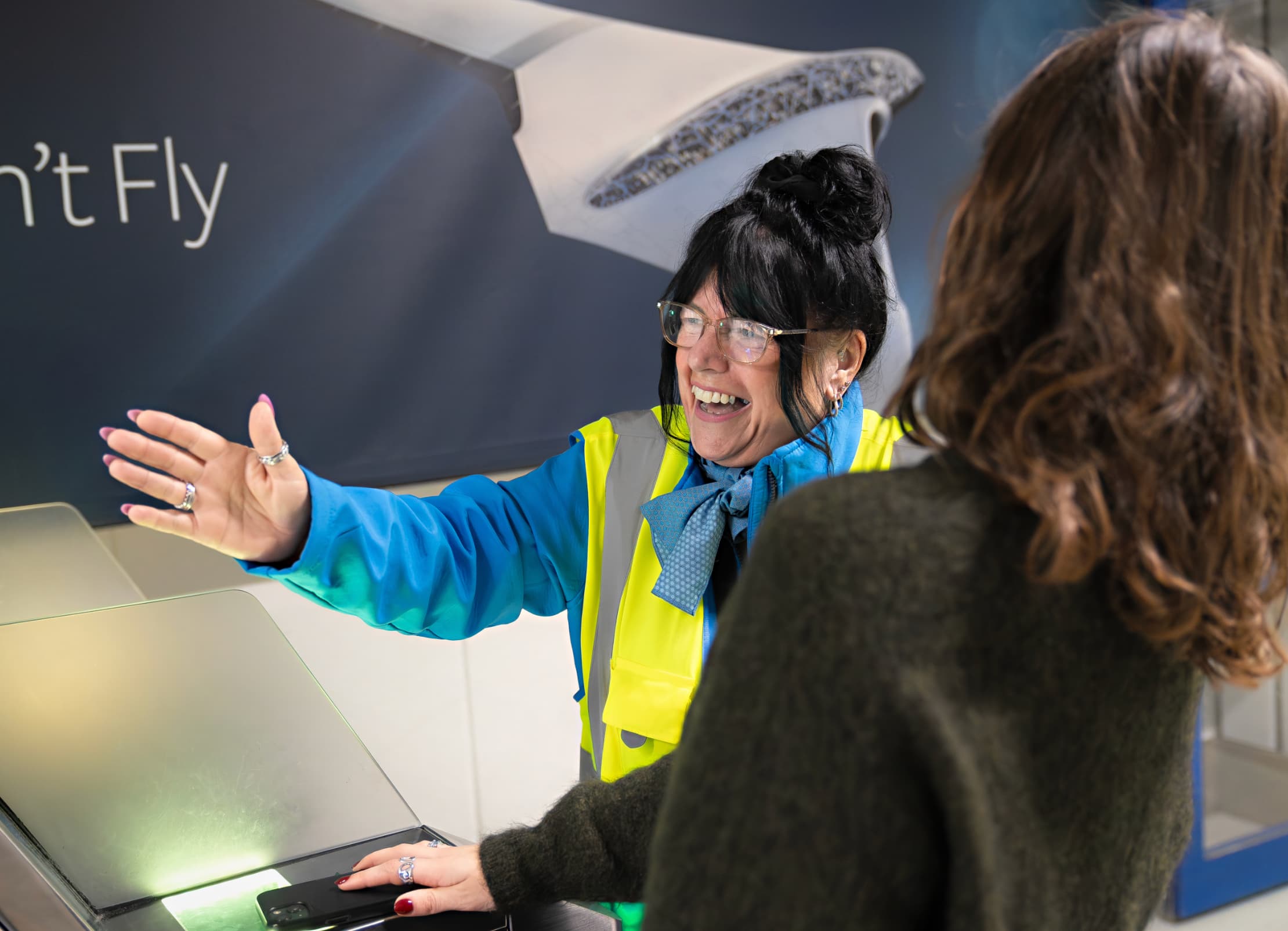 Smiling woman in high-vis vest and glasses gestures at another woman at a check-in counter.