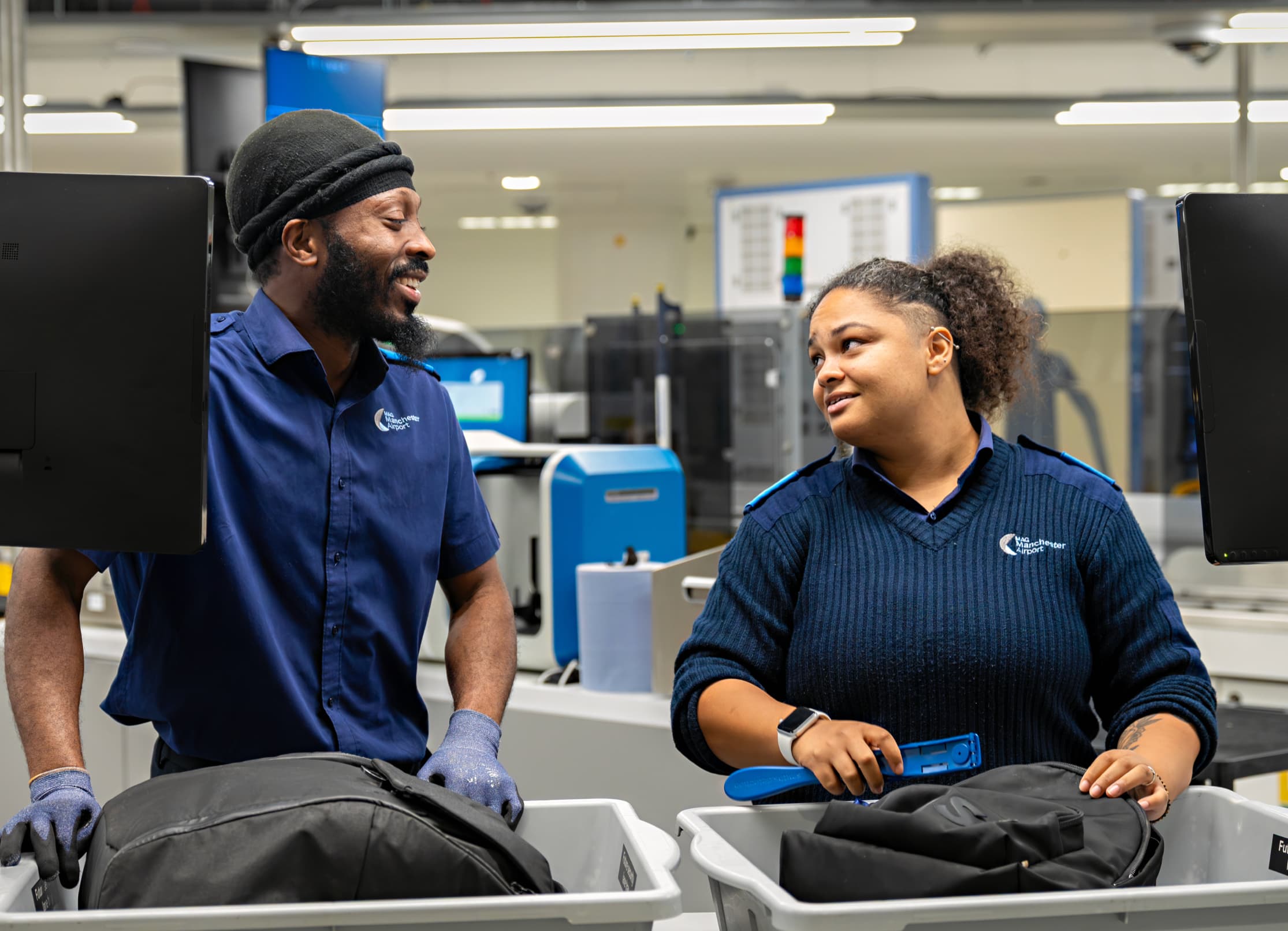 Two airport security staff in blue uniforms are smiling and talking while examining bags at a security checkpoint.