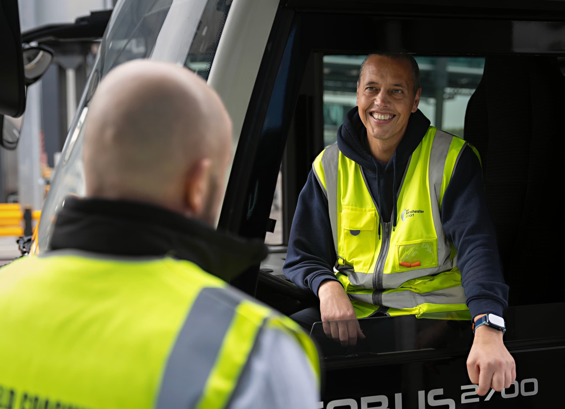 Two workers in high-visibility vests converse, one seated in a vehicle, smiling at the other who is standing outside.