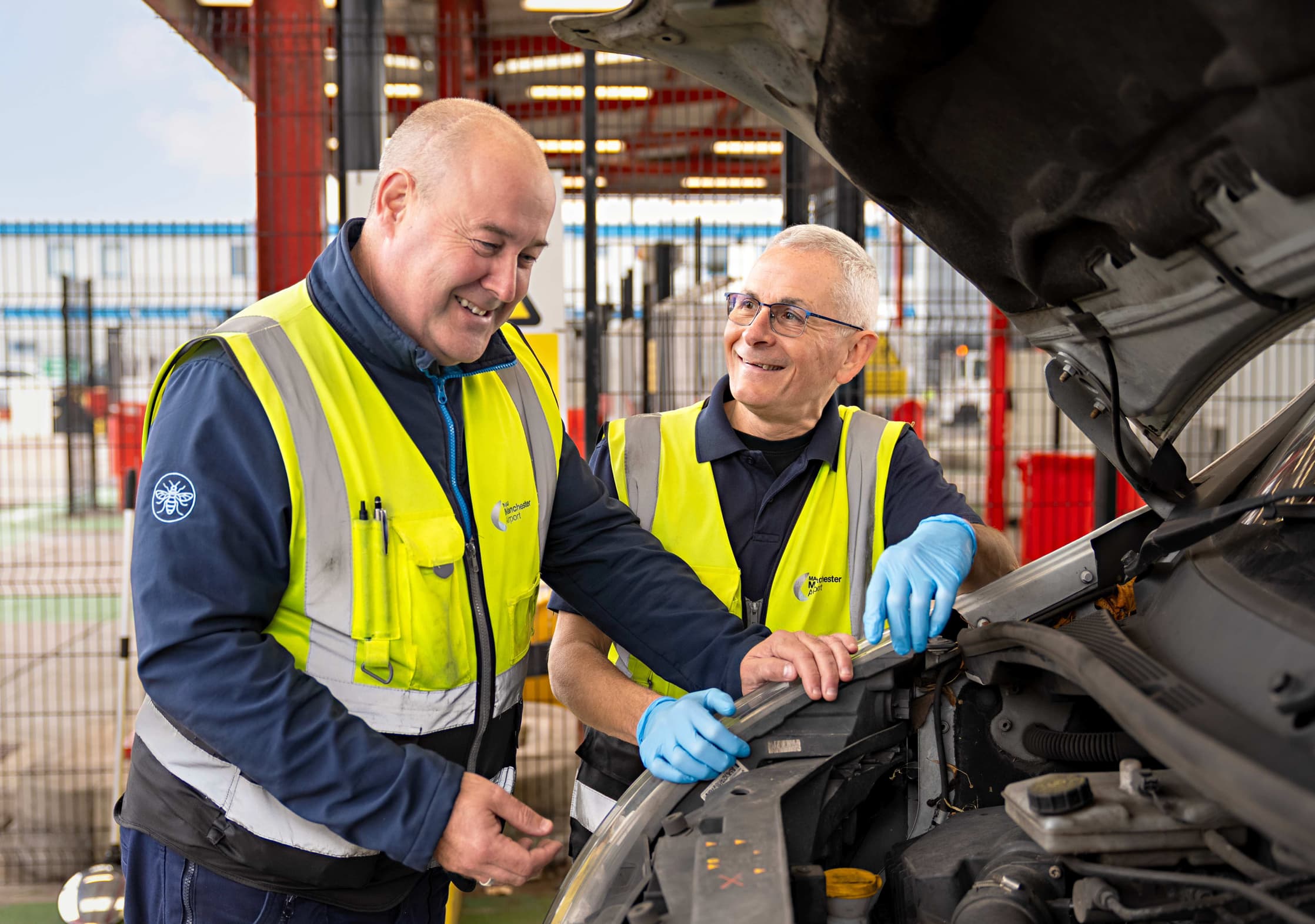 Two men in high-visibility vests and gloves work together, inspecting a car engine, smiling in a garage setting.