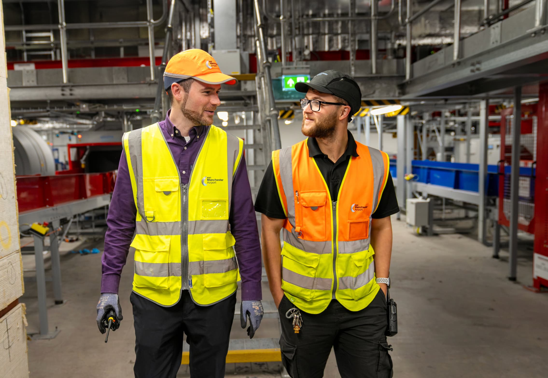 Two workers in high-visibility vests and hats walk together in an industrial setting, engaging in conversation.