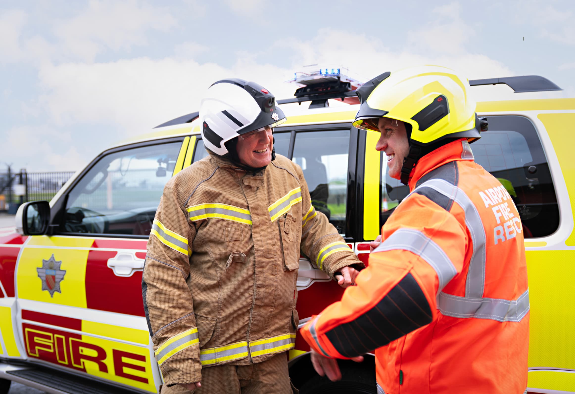 Two firefighters in protective gear converse cheerfully beside a fire rescue vehicle, with one wearing an "Airport Fire Rescue" jacket.