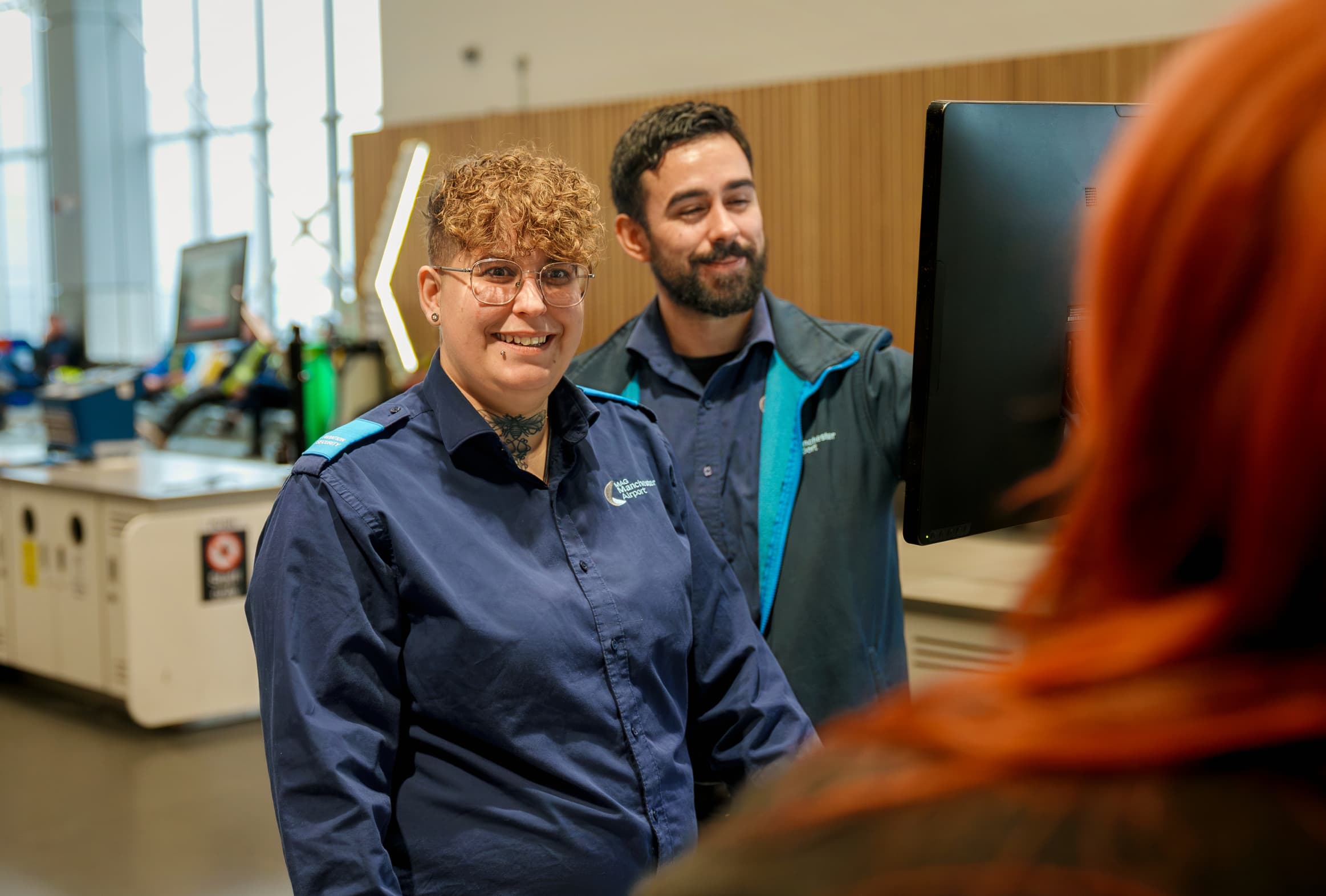 Two uniformed airport staff smiling and looking at a computer screen, assisting a person with red hair in a terminal.