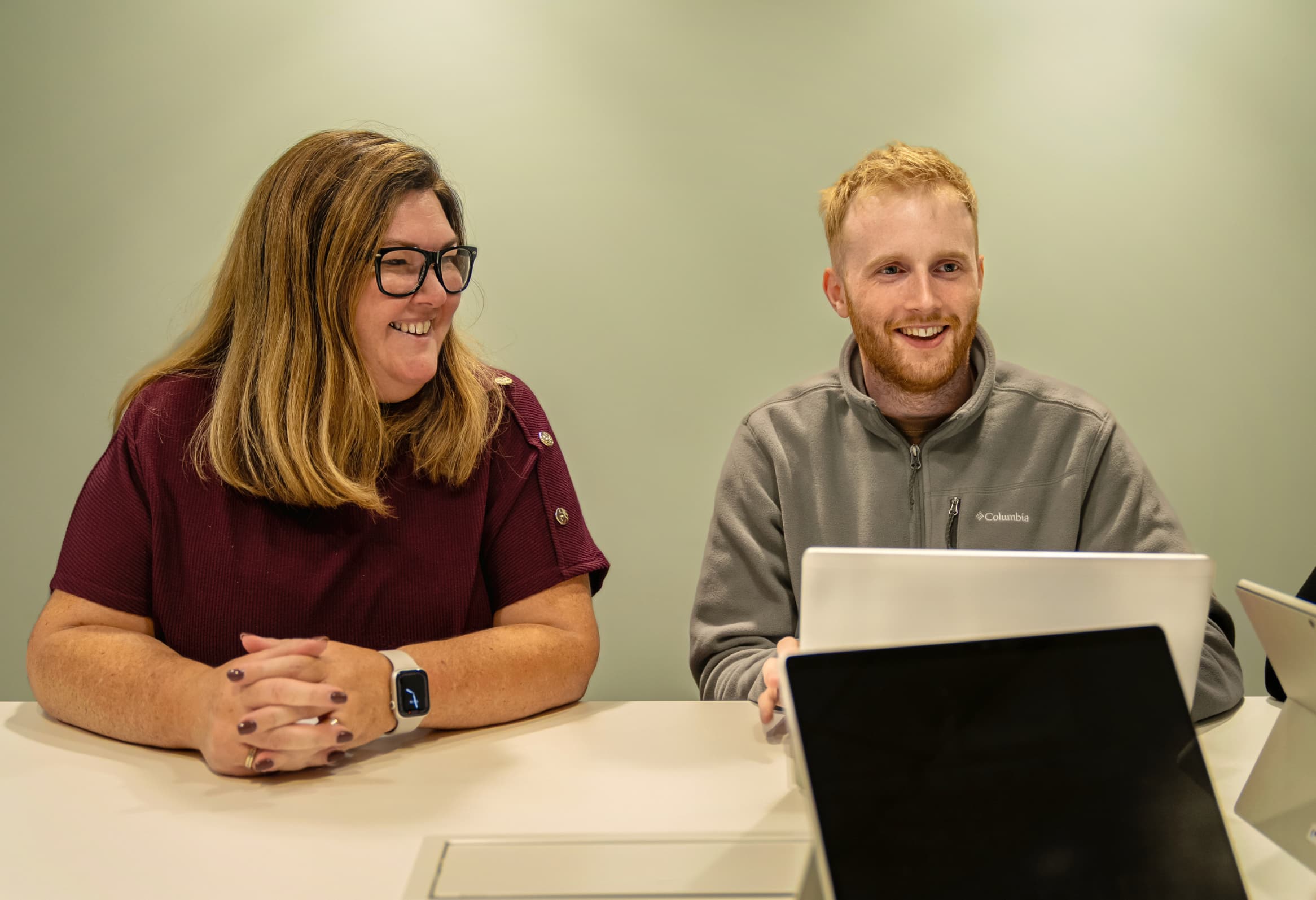 Two people smile while sitting at a table with laptops, engaged in a conversation or work session.