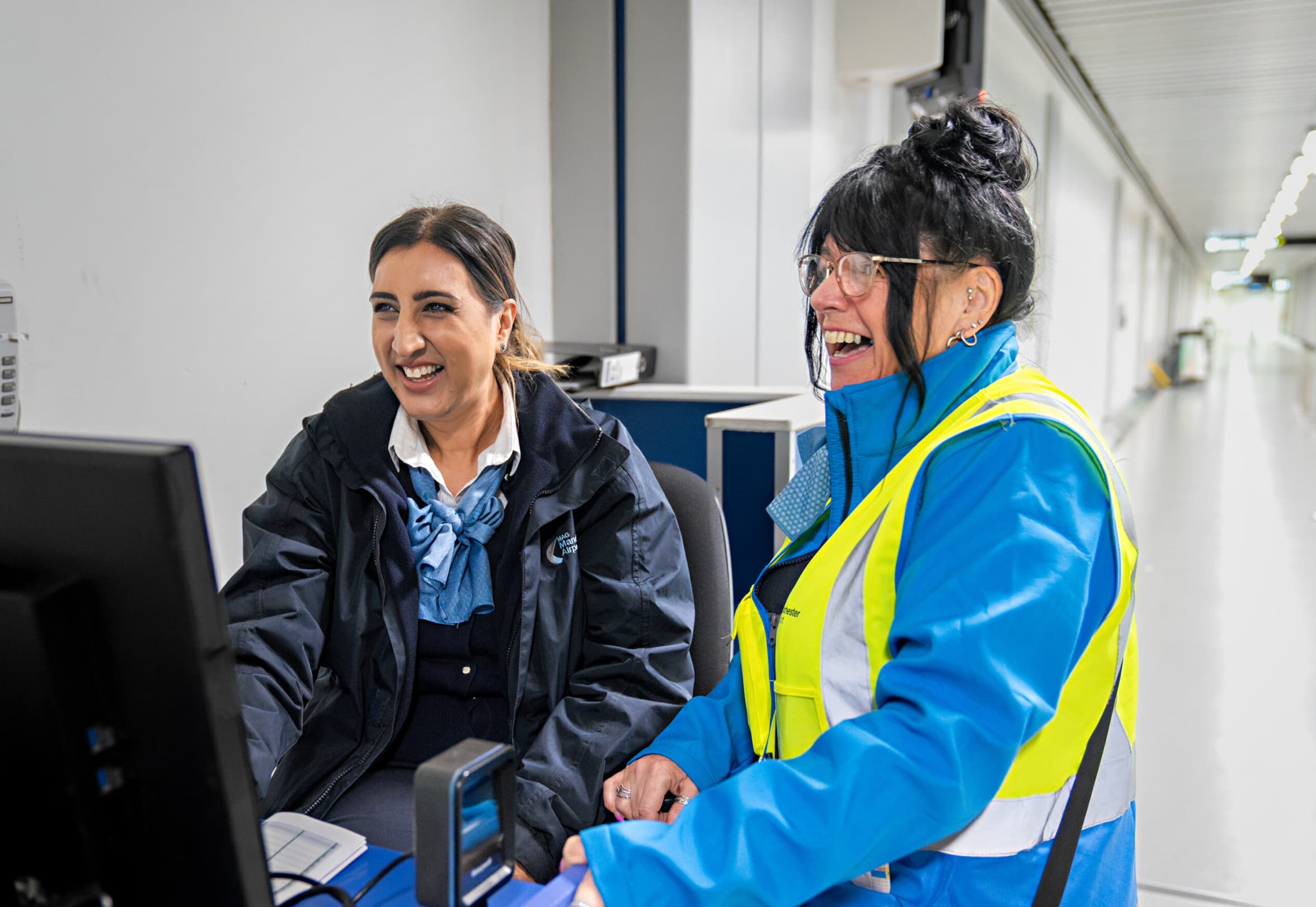 Two women in uniform laugh while sitting at a desk with a computer and scanner, in a brightly lit office corridor.
