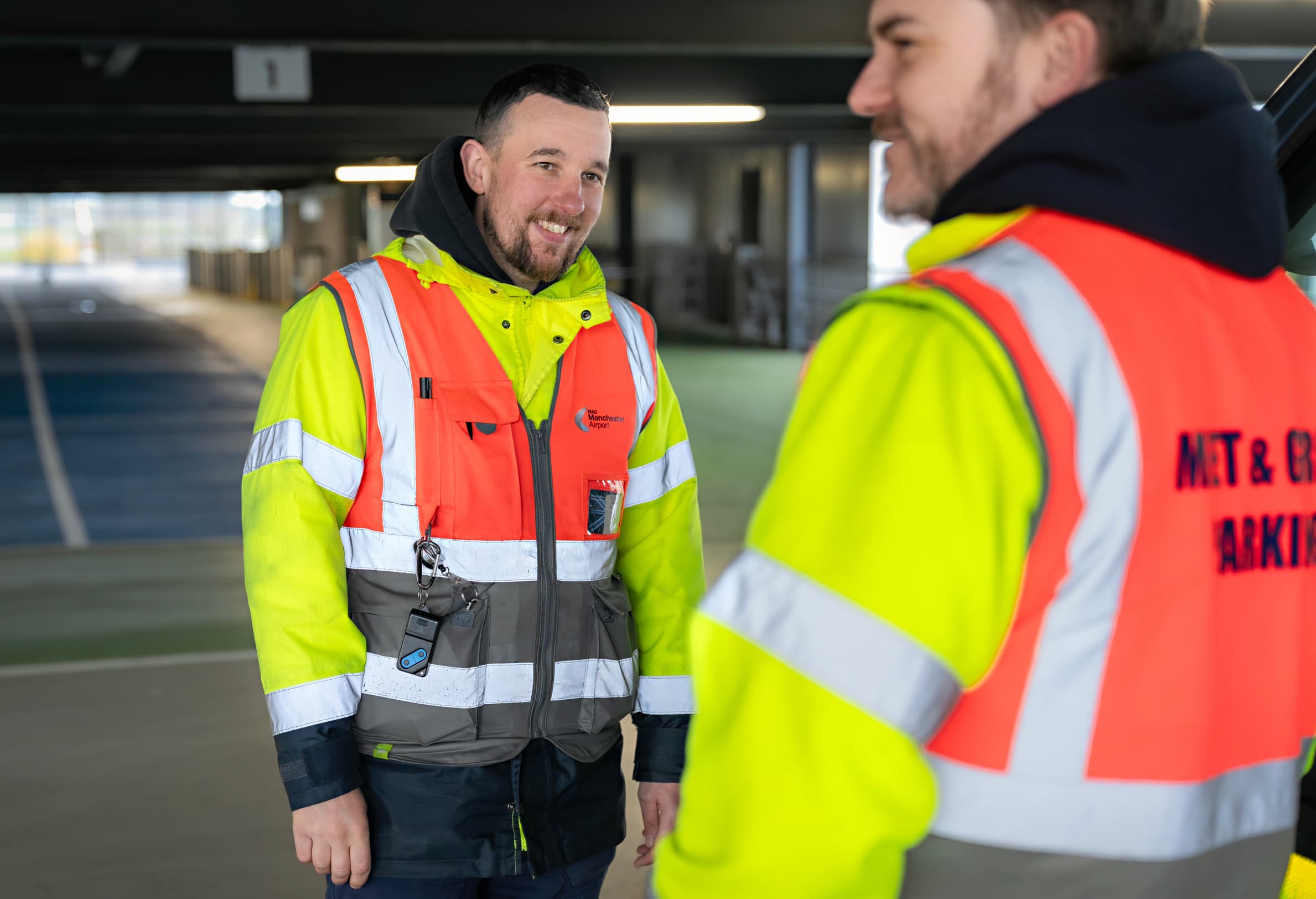 Two workers in reflective vests converse in a parking garage, one smiling, with keys and a device clipped to his vest.