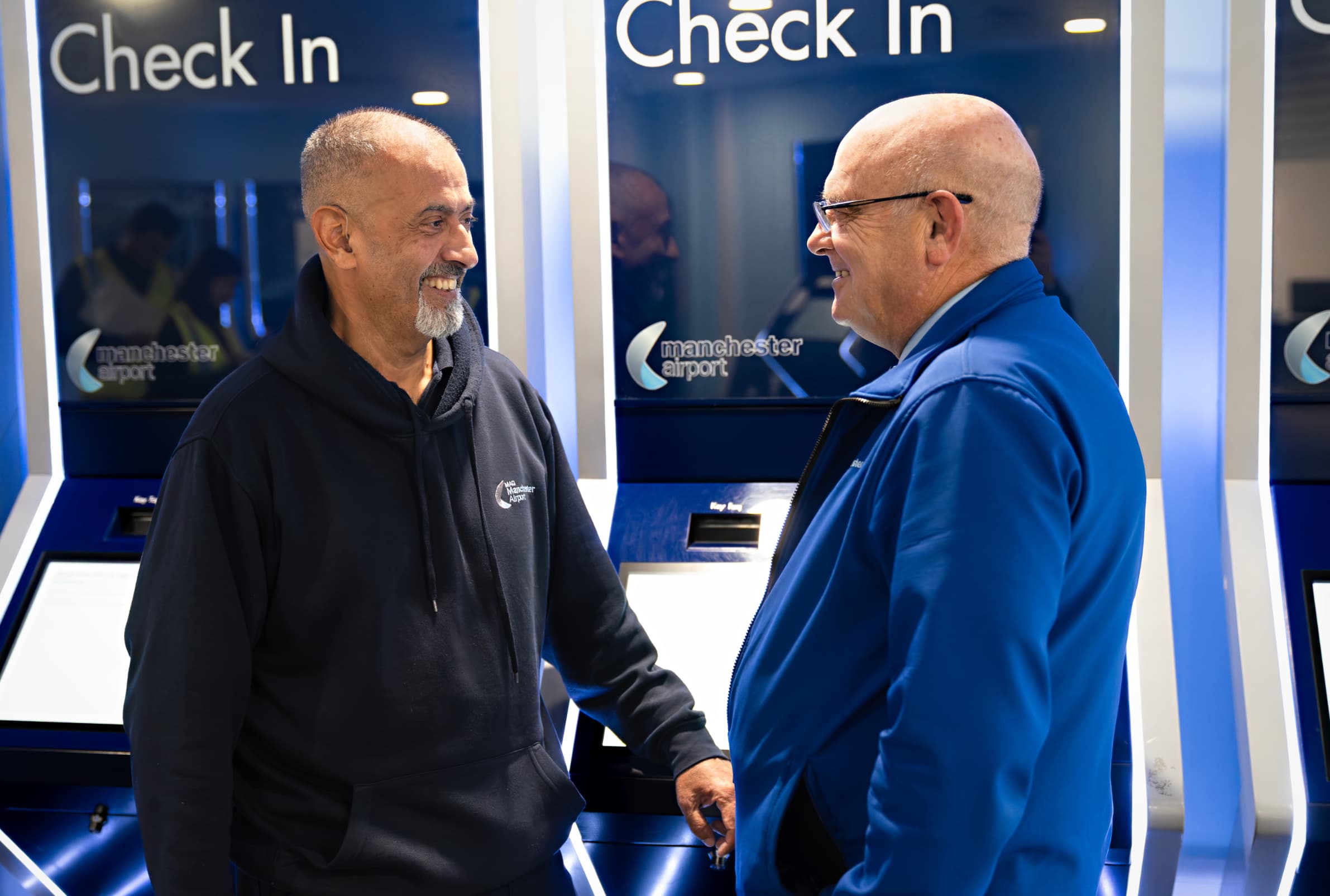 Two men smiling at each other at a Manchester Airport check-in area. One wears a black hoodie, the other a blue jacket.