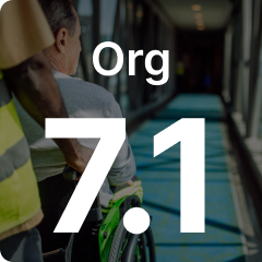 A person in a yellow vest assists another in a wheelchair down a long corridor with "Org 7.1" text overlay.