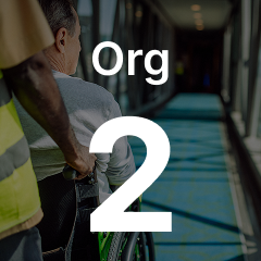 A person in a wheelchair is assisted by another on a long corridor with "Org 2" written over the image.