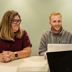 Two people sitting at a table with laptops, engaged in conversation, smiling, and appearing relaxed and happy.