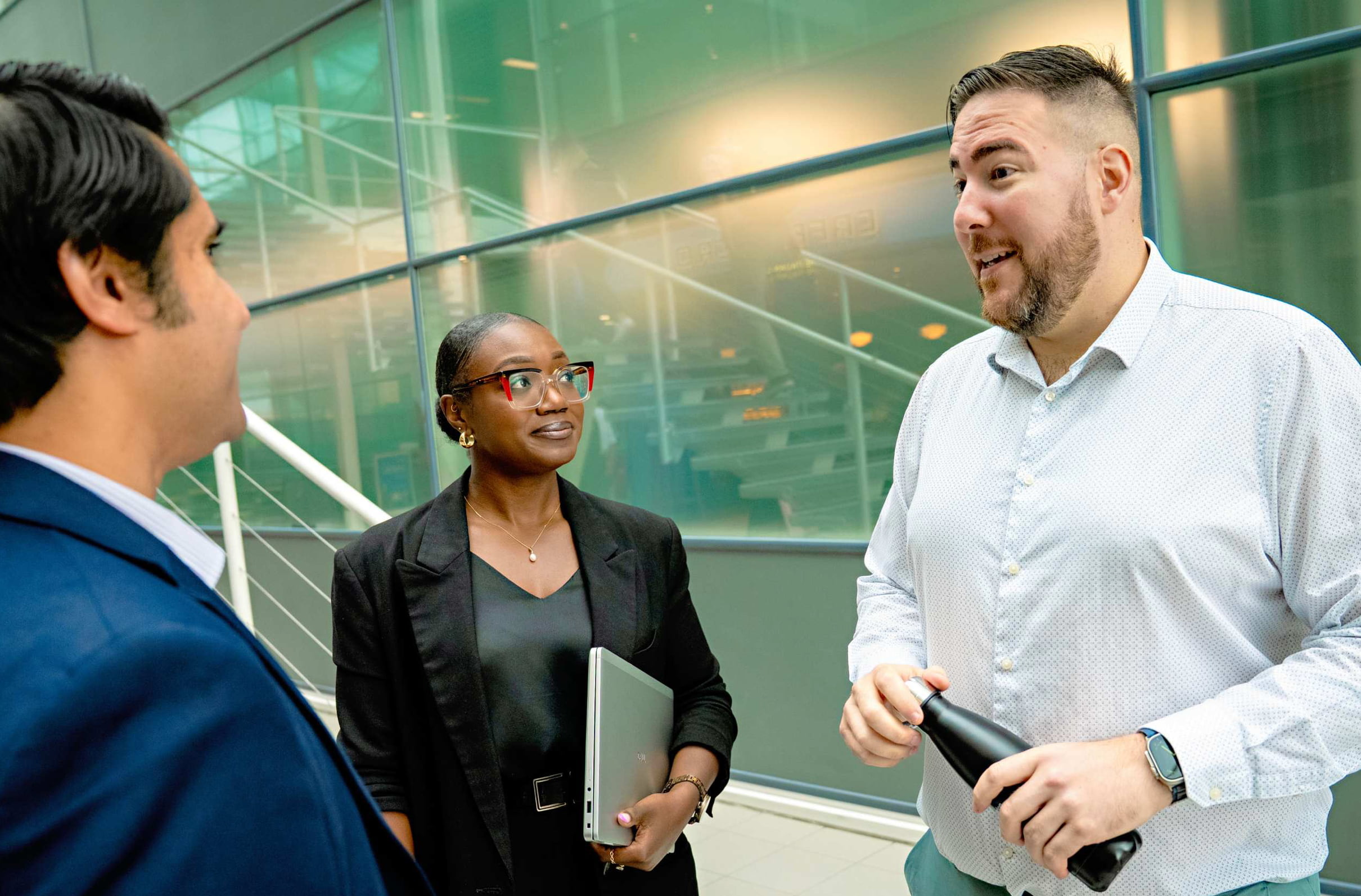 Three people engaged in conversation in a modern office, with one holding a laptop and another with a drink bottle.