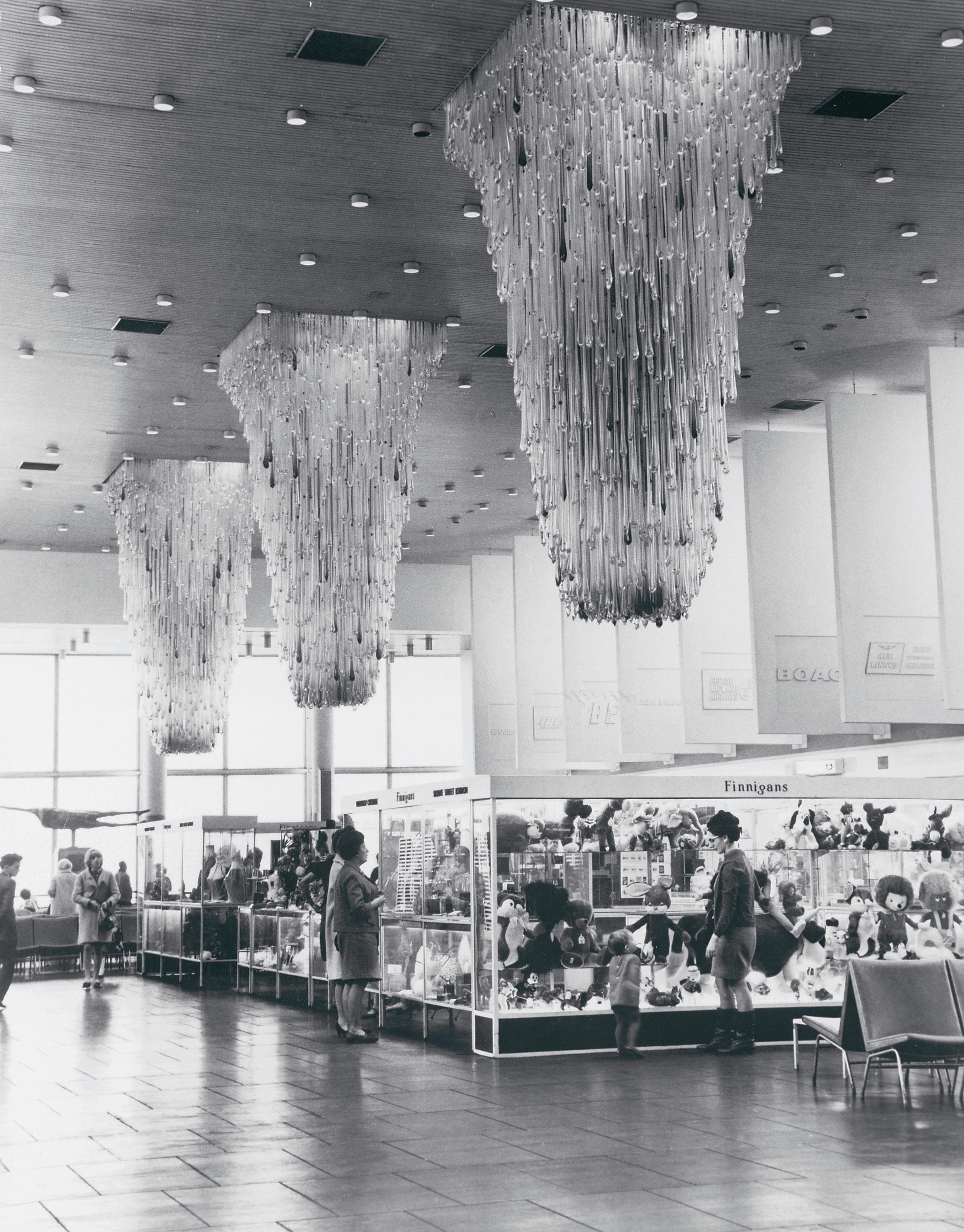 People browse display cases in a large room with elaborate chandeliers hanging from the ceiling, creating a striking pattern above.