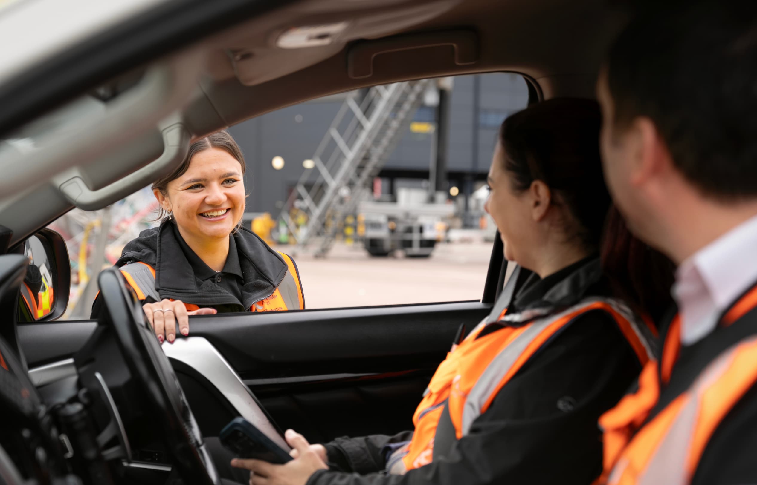 Smiling worker in a reflective vest talks with two colleagues in a car, parked at an industrial site.