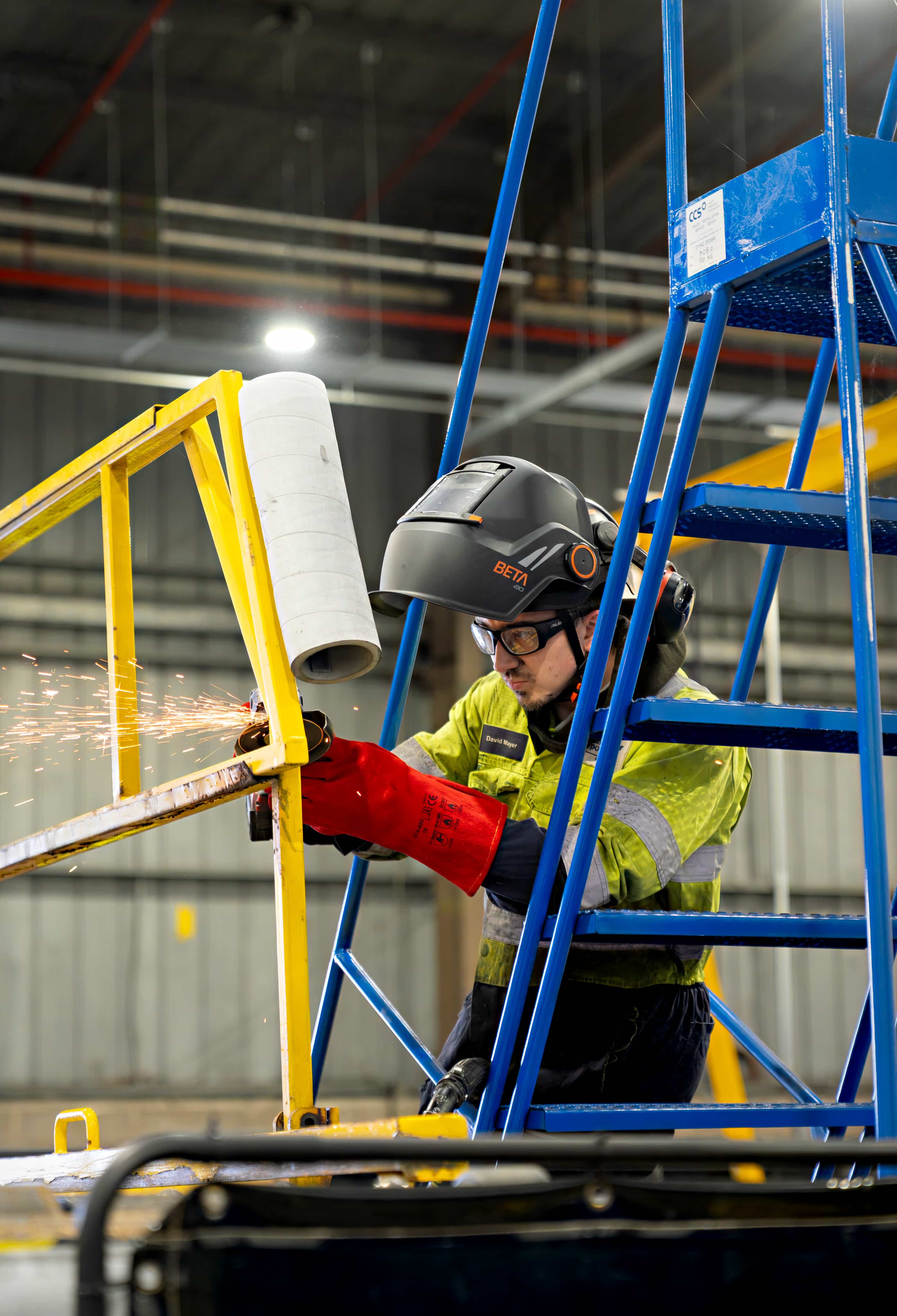 A worker in protective gear welds metal, producing sparks, while standing on blue stairs in an industrial setting.