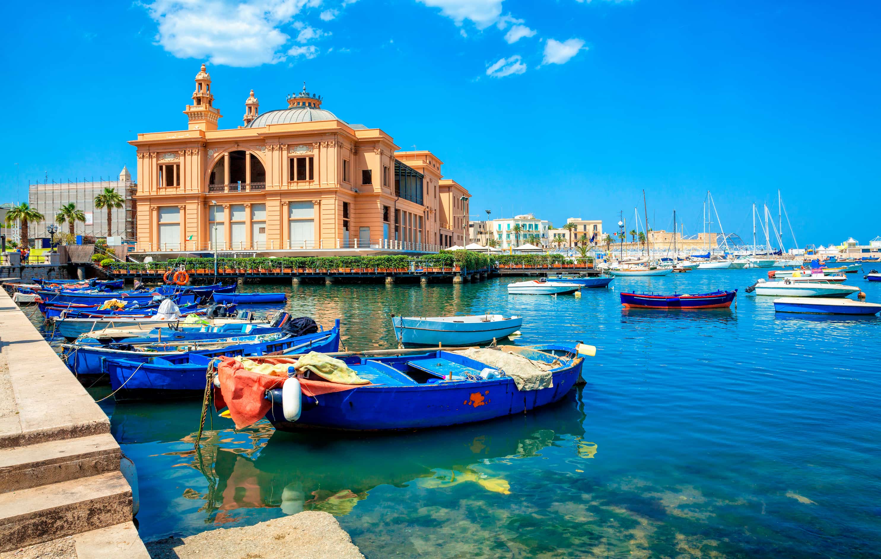 Colourful boats docked in clear blue water near a grand historic building under a bright blue sky.