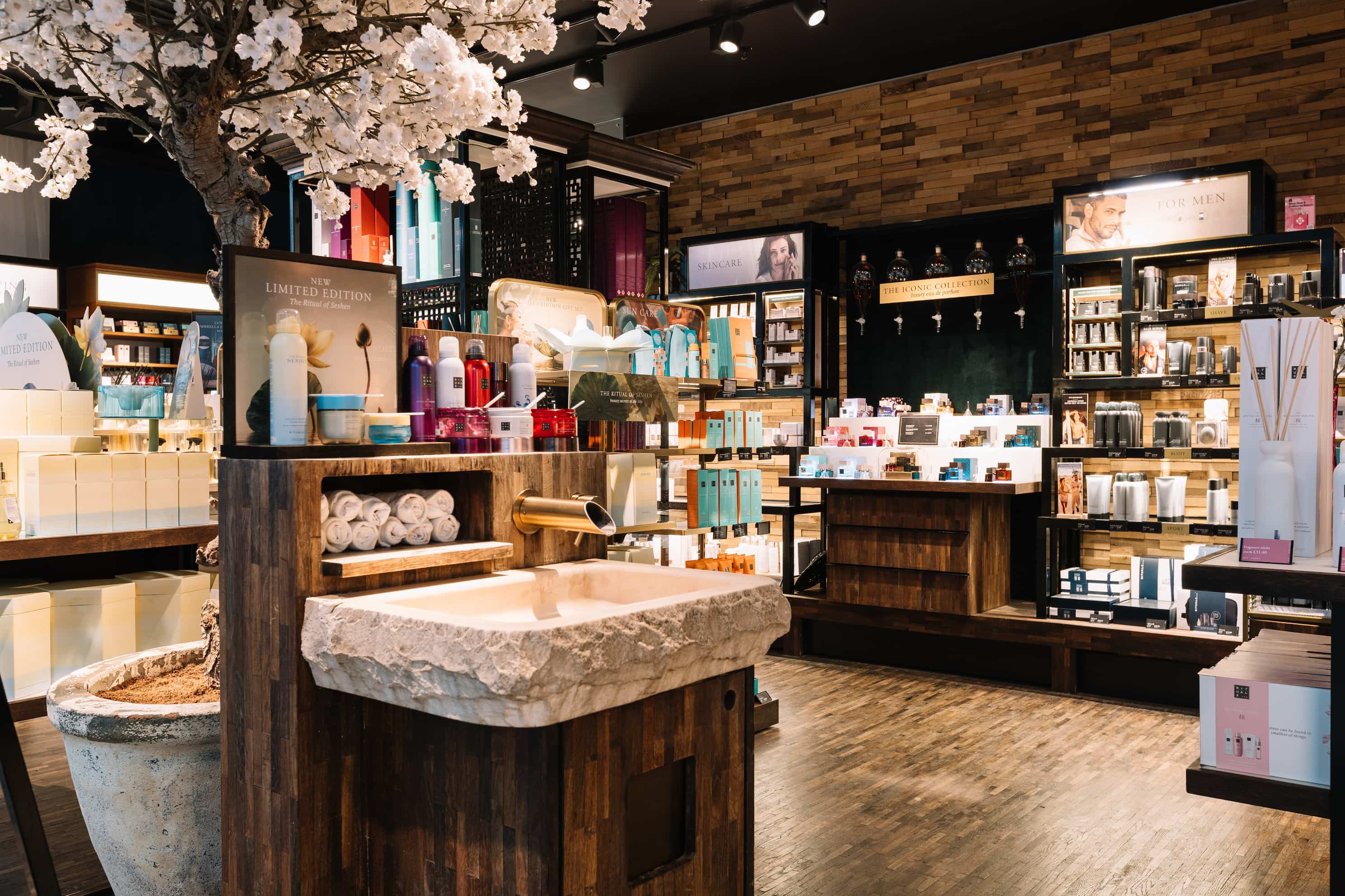 Luxurious skincare store with wooden décor, a stone sink, flower arrangements, and well-organised shelves of products and towels.