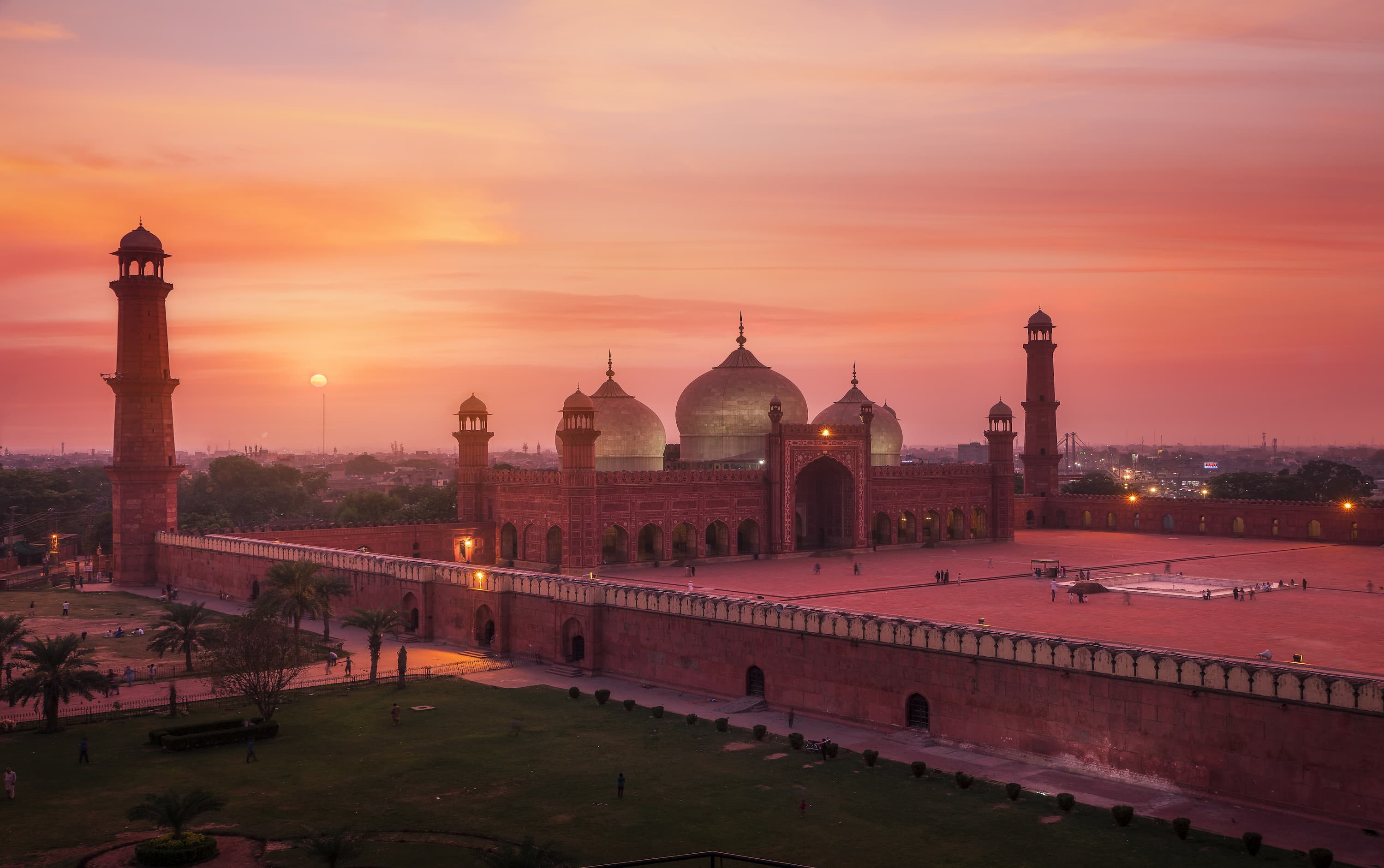 A large mosque with three domes at sunset, surrounded by tall minarets and an expansive courtyard, silhouetted against a vibrant orange sky.