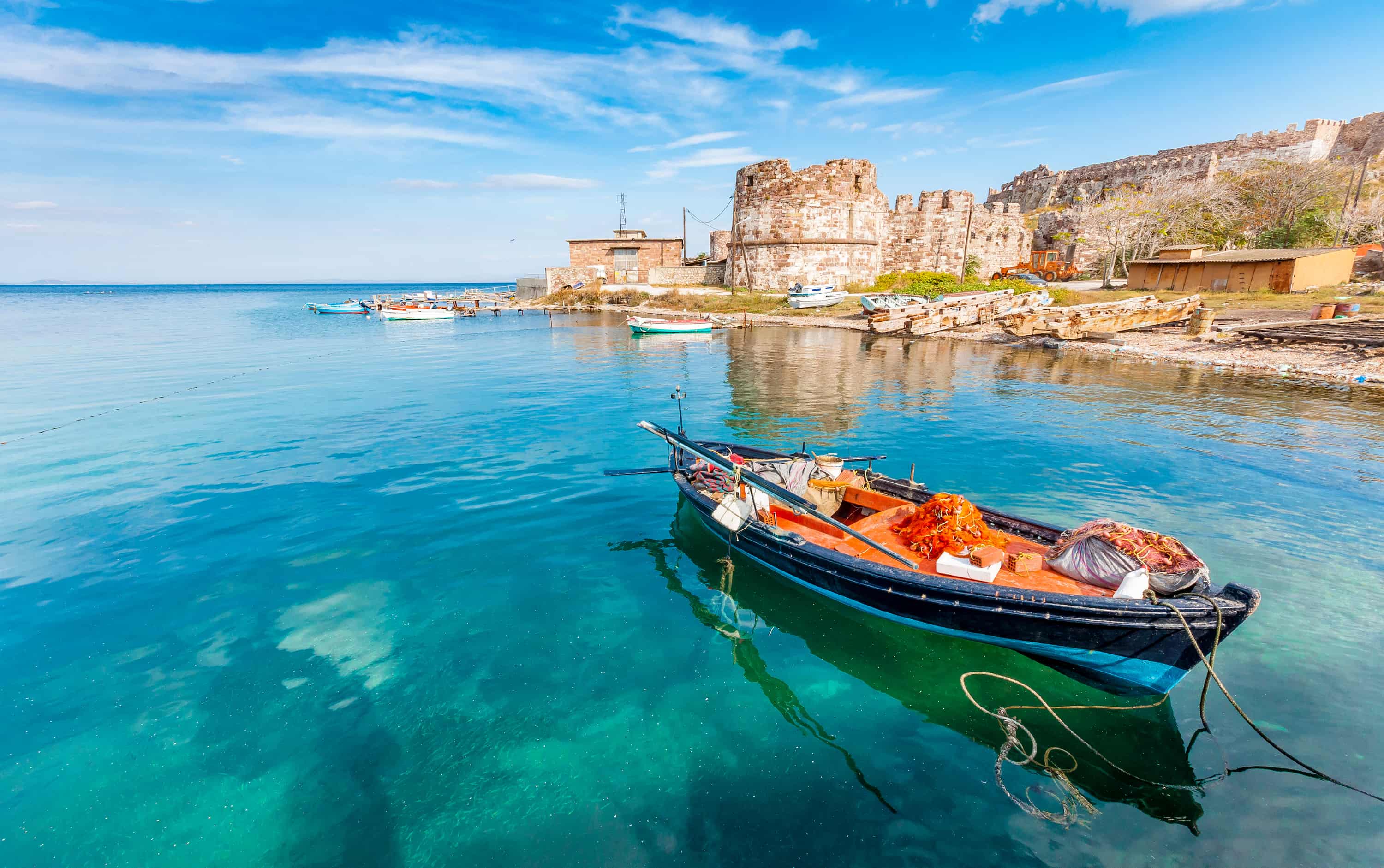 A small fishing boat floats in clear turquoise water near a rustic stone fortress on a bright day with blue skies.