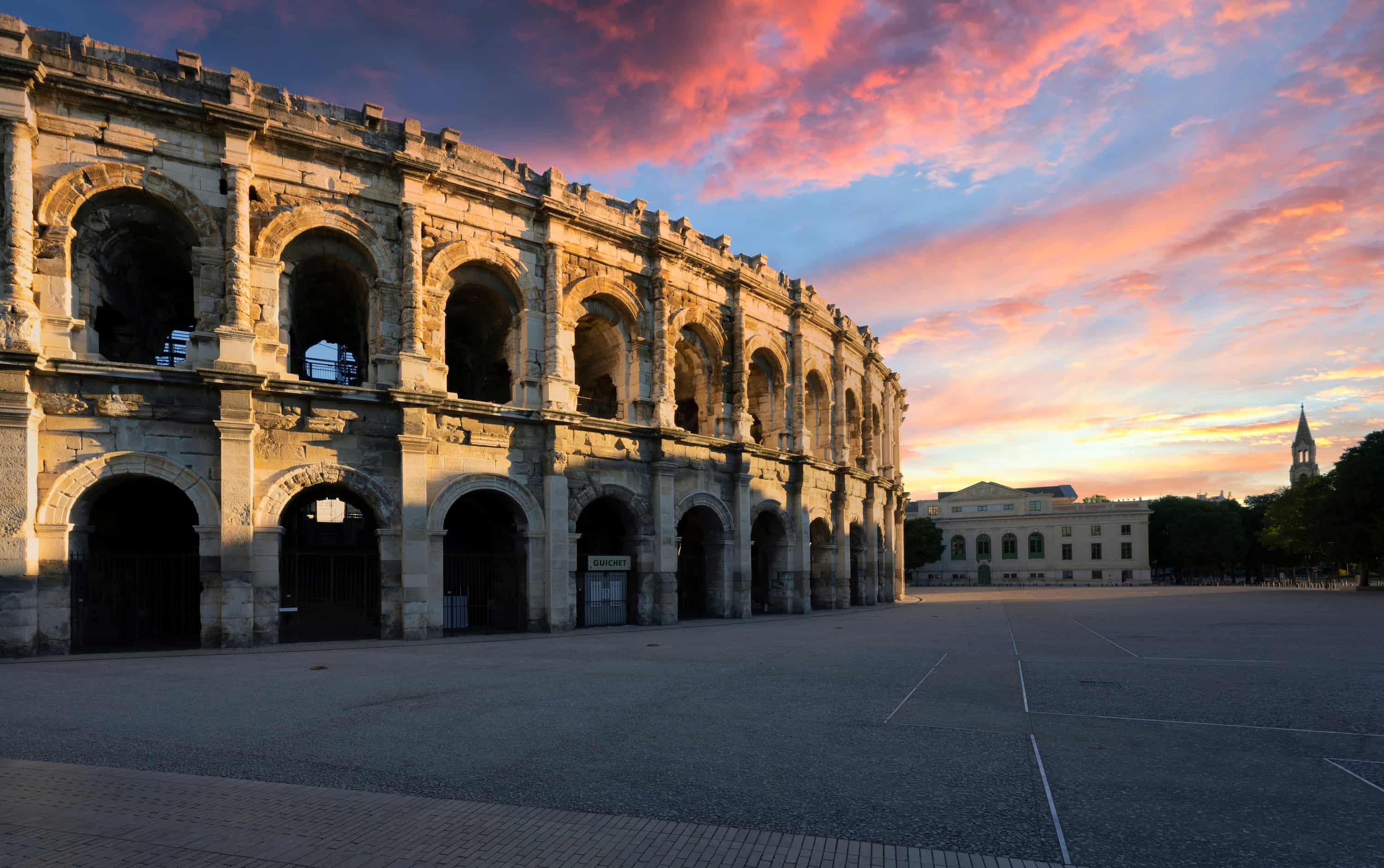 Ancient Roman amphitheater under a colorful sunset sky, with a spire and building in the background, set in an open plaza.