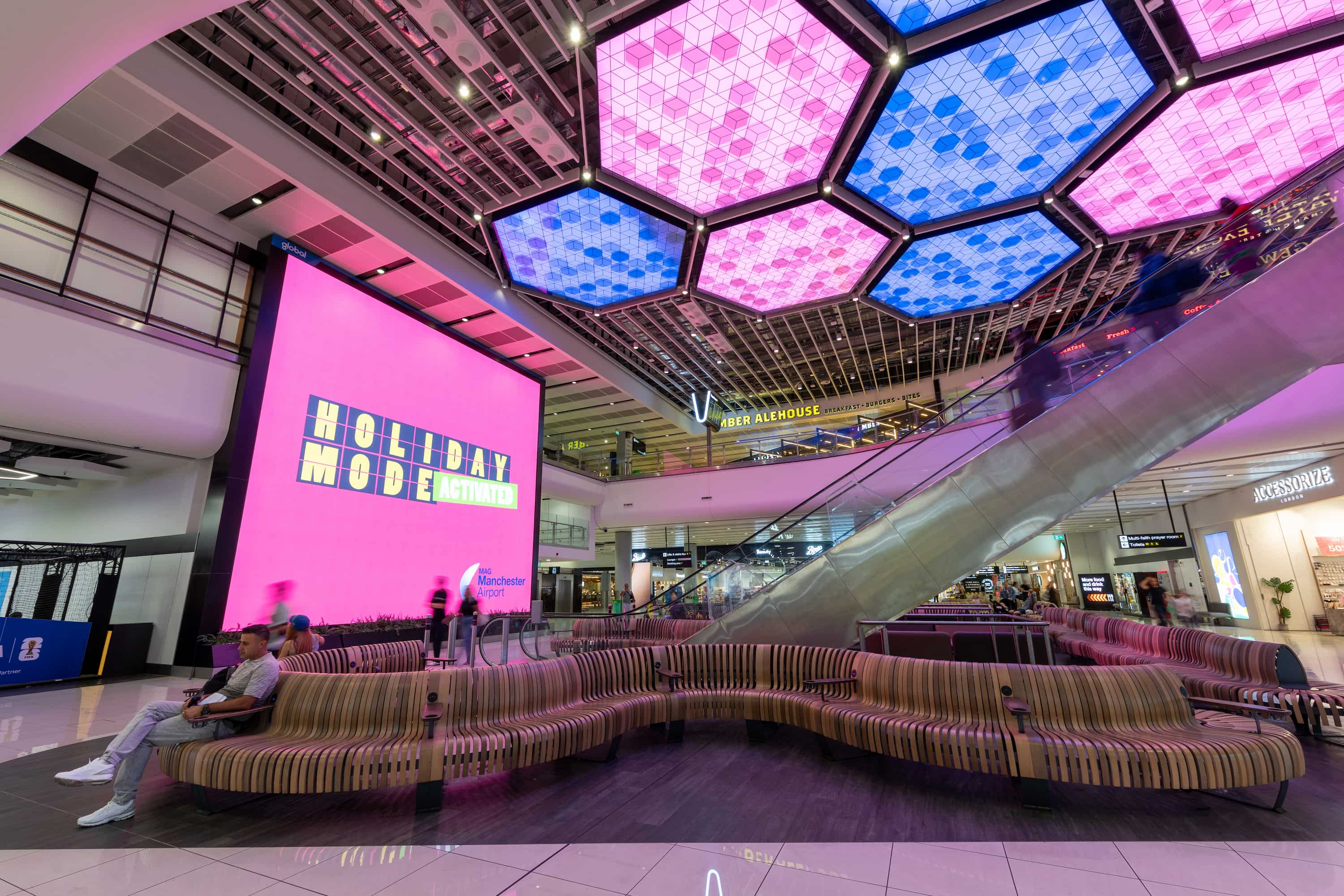 Colourful airport interior with hexagon-patterned ceiling lights, a pink "Holiday Mode Activated" sign, escalator, and seating area.