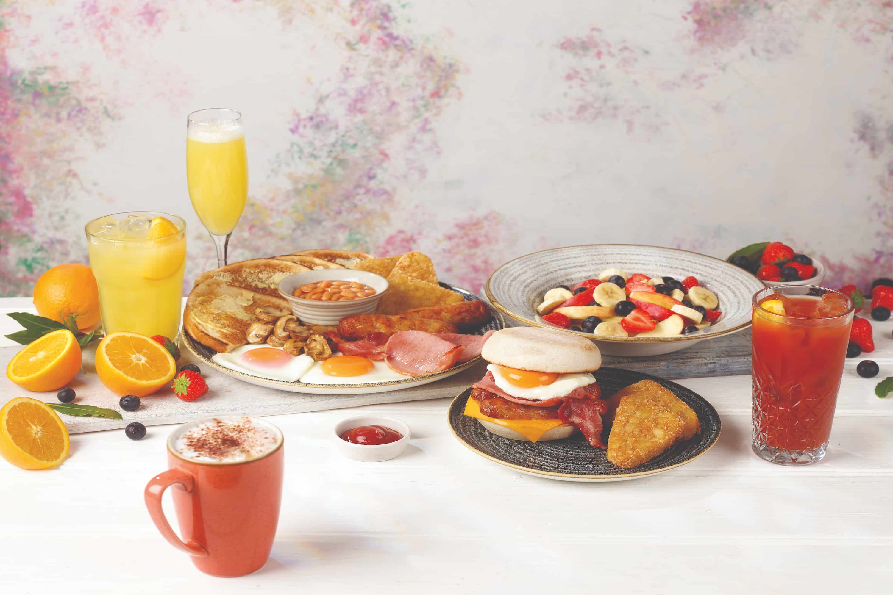 A variety of breakfast foods including eggs, pancakes, fruit, an English muffin sandwich, hash brown, coffee, and juices displayed on a table.