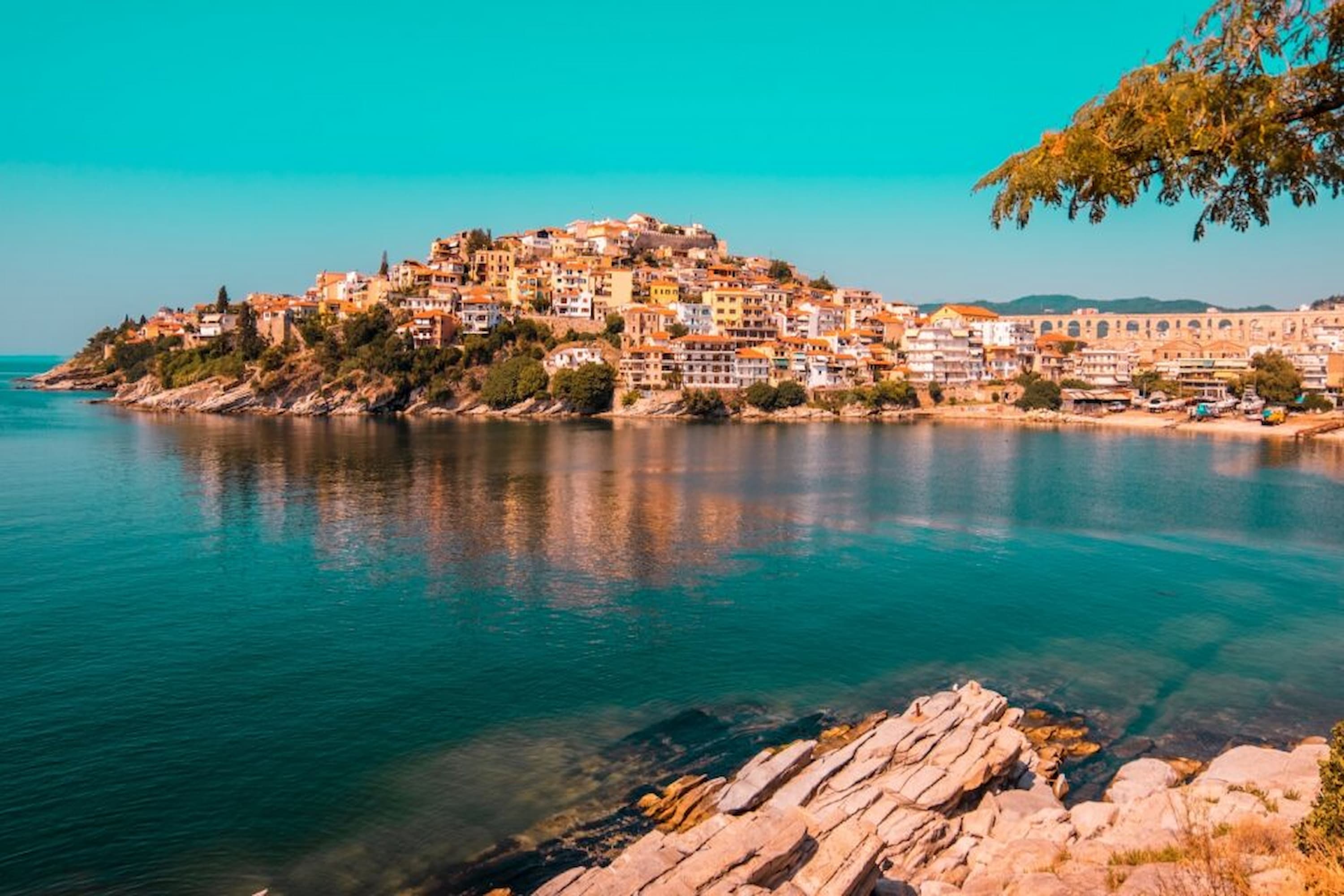 A coastal town with colourful buildings atop a hill overlooks a calm, blue sea under a clear sky, with rocks and trees in the foreground.