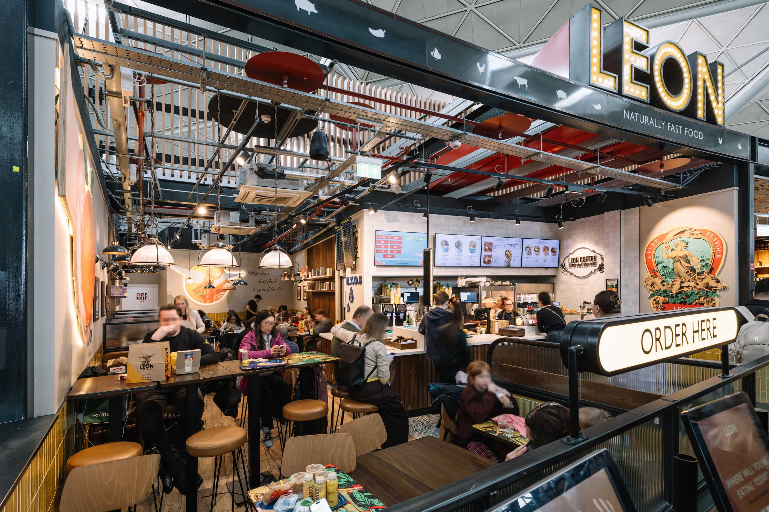 Interior of a bustling fast-food restaurant with passengers dining and ordering at the counter under a sign reading "LEON."