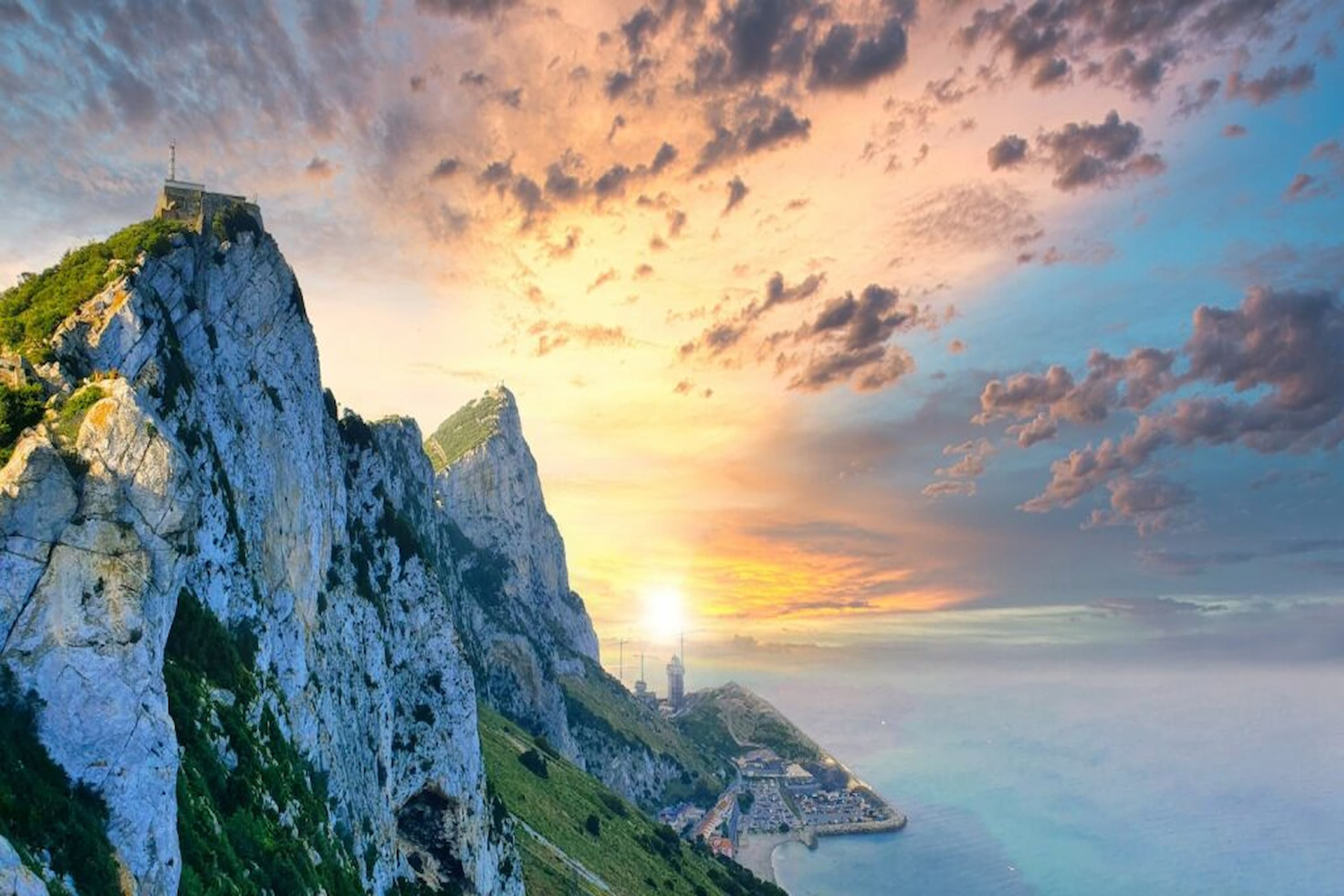 Sunset view of the Rock of Gibraltar, with dramatic clouds above and calm sea and coastal landscape below.