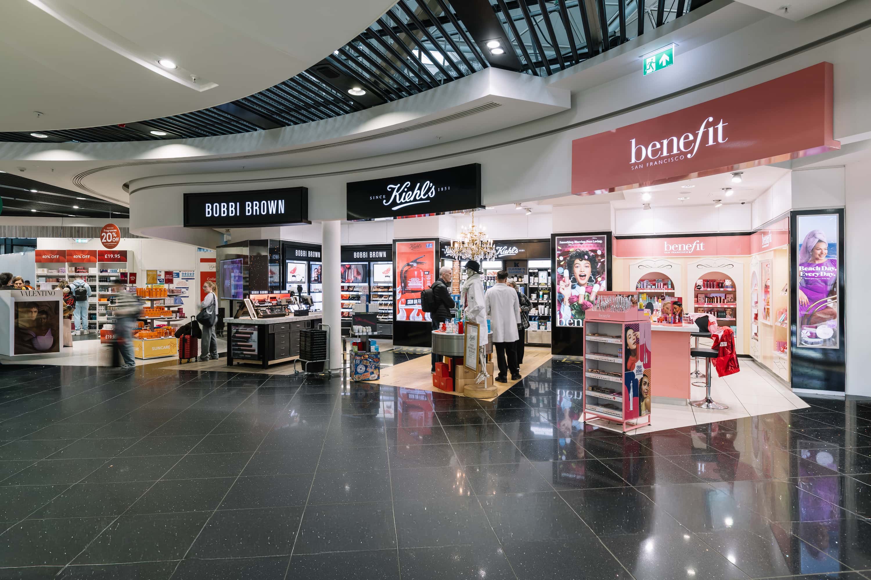 Cosmetics section in a mall with Bobbi Brown, Kiehl’s, and Benefit stores, featuring makeup displays and shoppers browsing products.