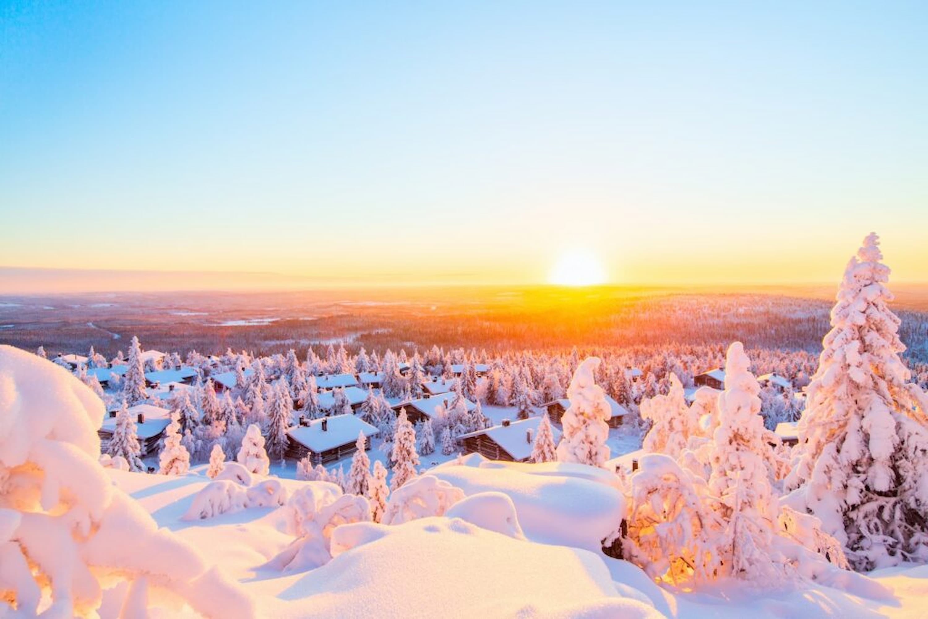 Snow-covered trees and cabins under a vibrant, golden sunset on a clear day, with a sprawling snowy landscape in the background.