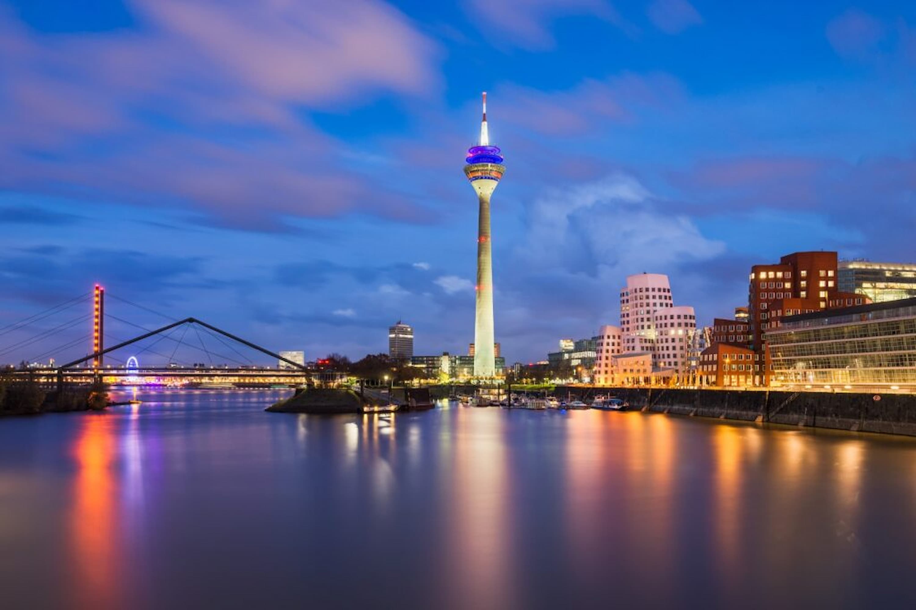 Cityscape at dusk featuring a tall illuminated tower by the river, with modern buildings and a bridge in the background under a partly cloudy sky.