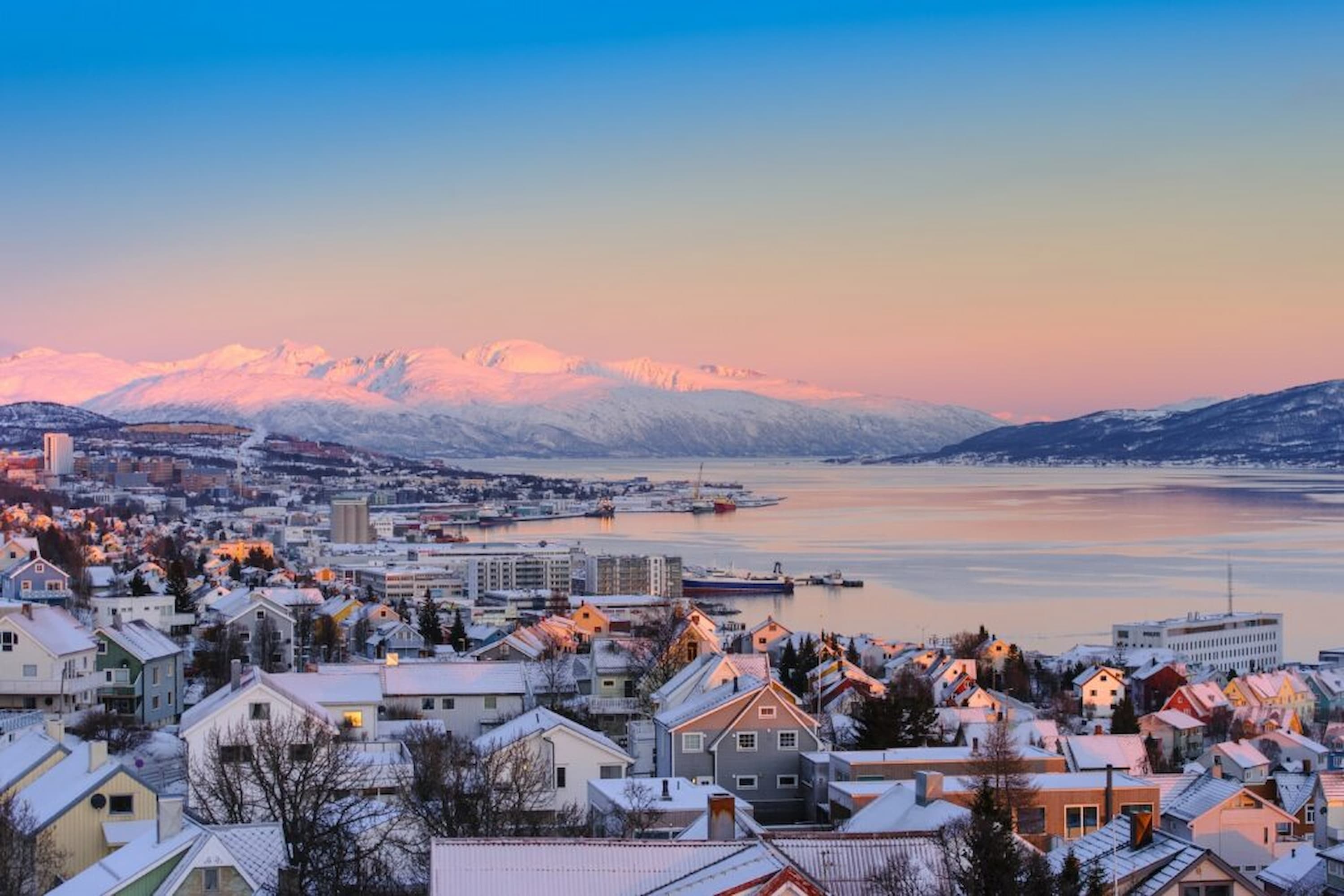 Snow-covered town with colourful houses by a calm fjord, backdropped by sunlit mountains under a pink and blue sky at sunrise.