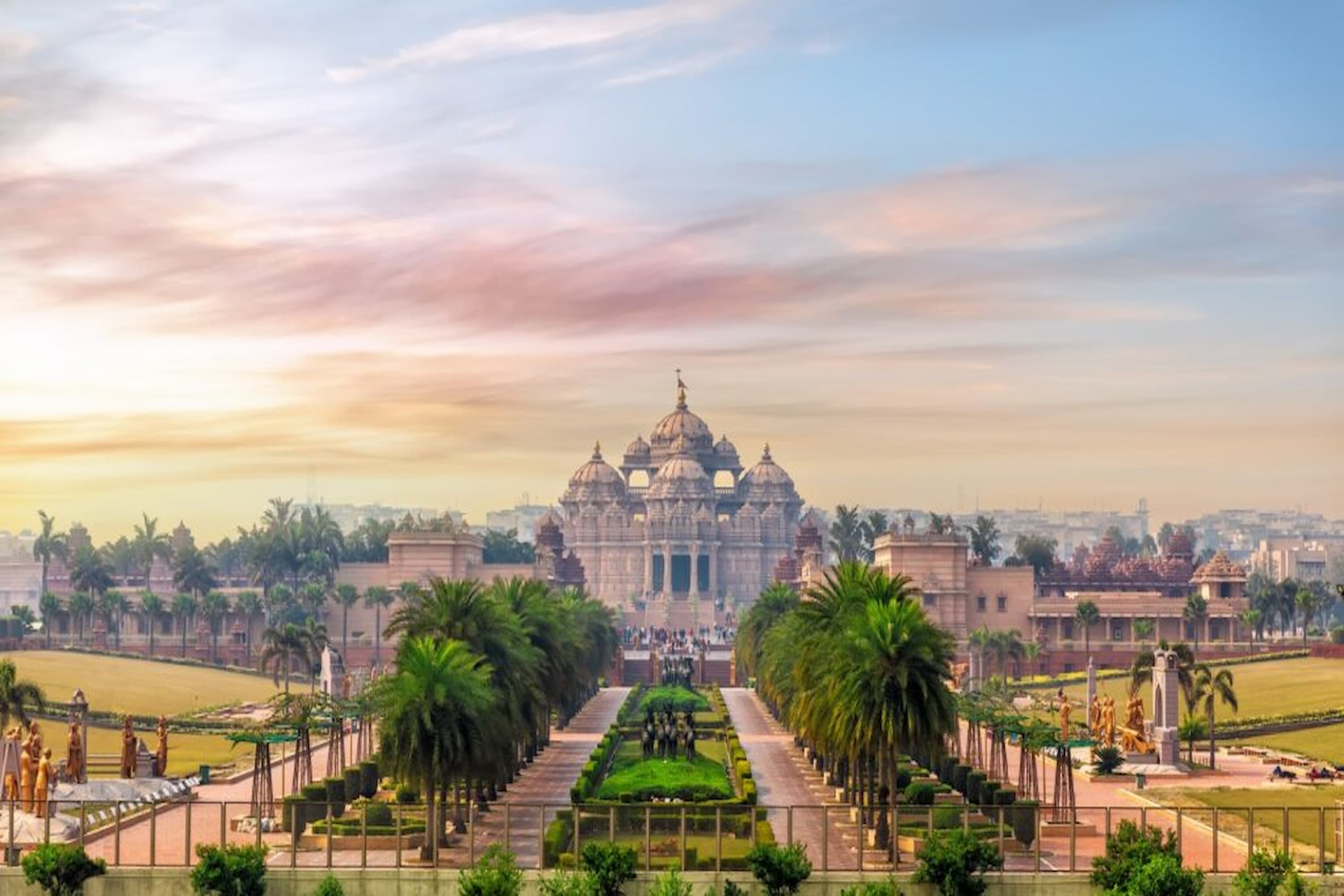 A grand temple complex with intricate architecture, surrounded by lush gardens and palm trees, under a colourful sky at sunset.