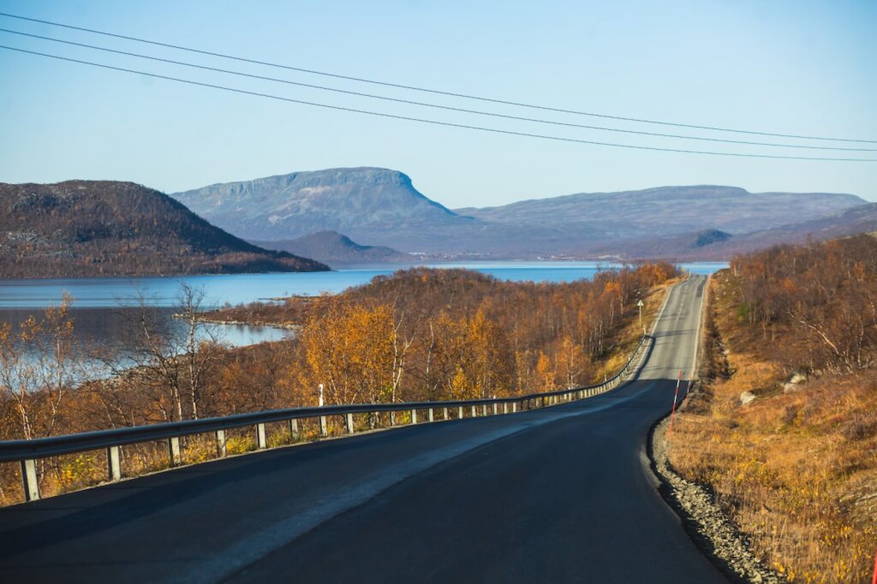 Scenic road with guardrails, flanked by autumn trees and hills, leads towards a tranquil lake and distant mountains under a clear blue sky.