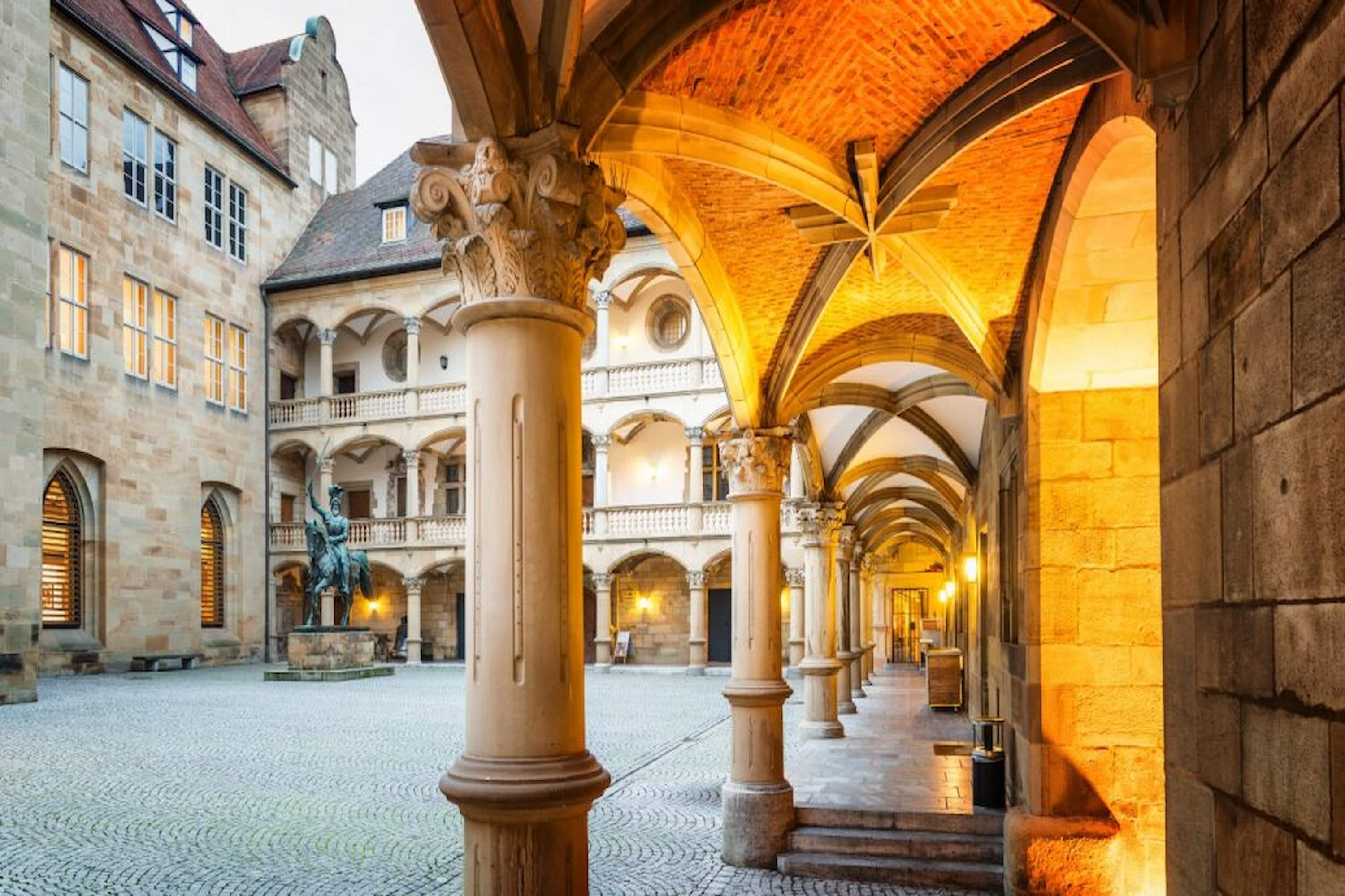 Historic courtyard with stone arches, columns, and warm lighting. Cobblestone ground and statue visible under a twilight sky.
