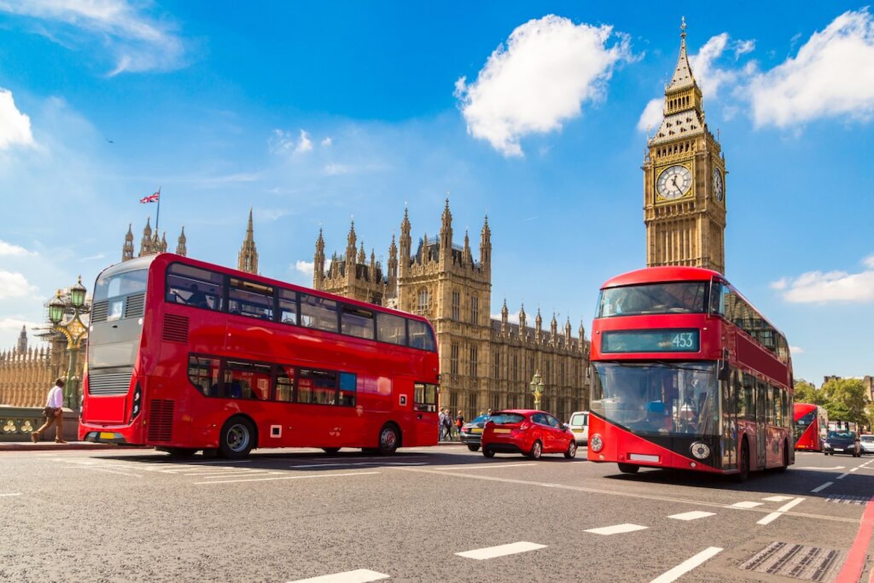 Red double-decker buses and cars on a road in front of Big Ben and the Houses of Parliament under a clear blue sky.
